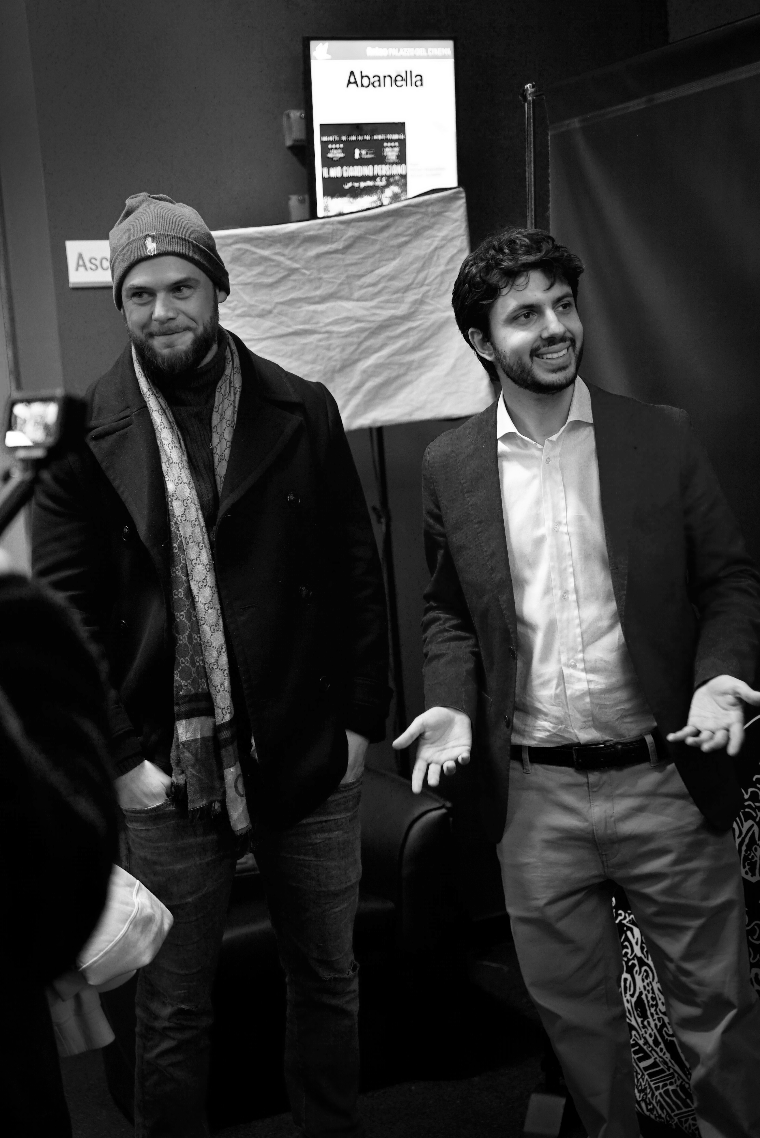 Two men posing, one wearing a beanie and scarf, the other in a suit, in a black and white setting.