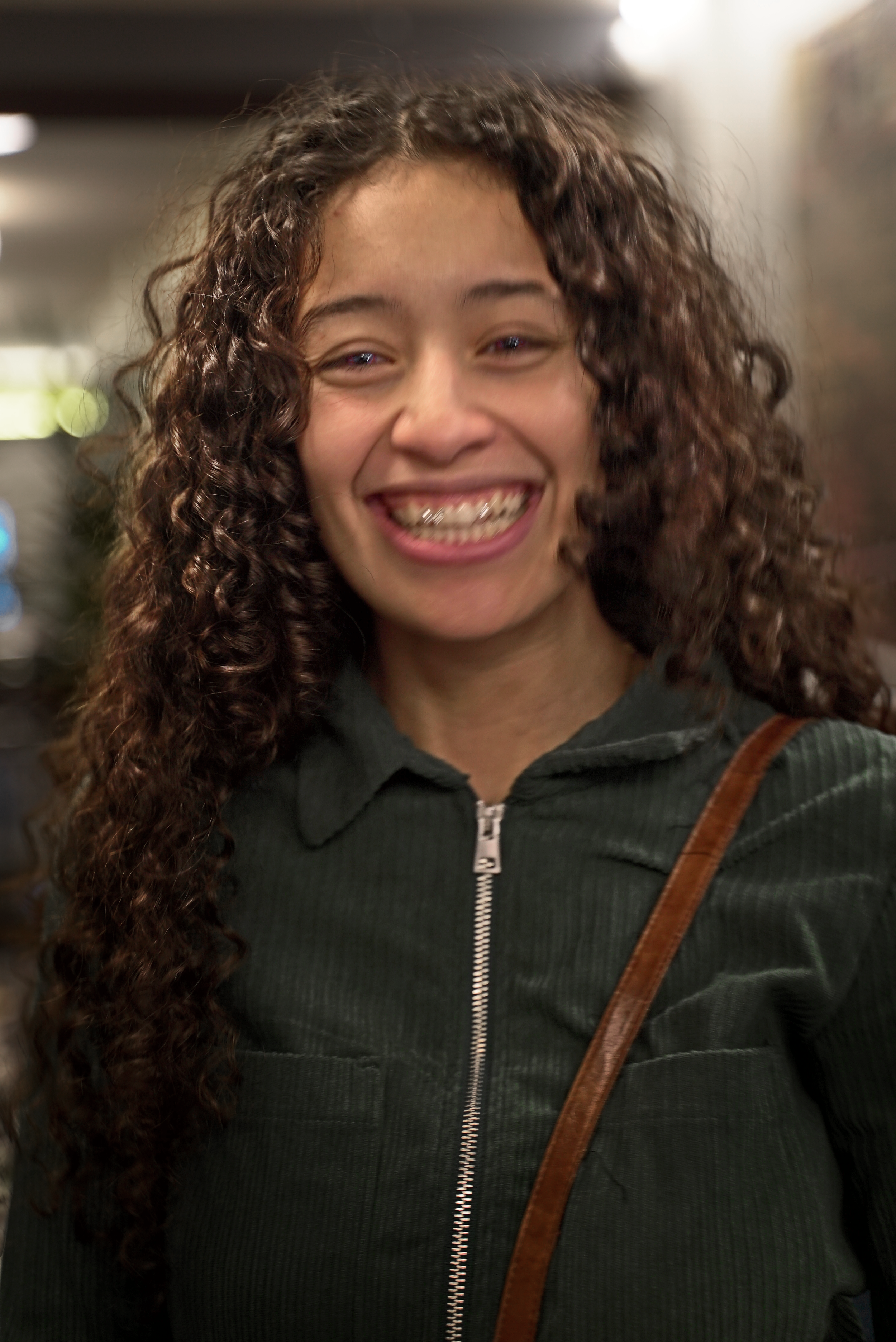 Young person smiling with curly hair and braces, wearing a dark green jacket indoors.