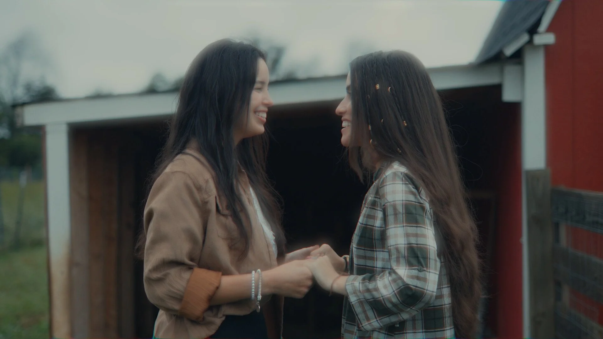 Two people smiling and holding hands outdoors, standing in front of a wooden structure.