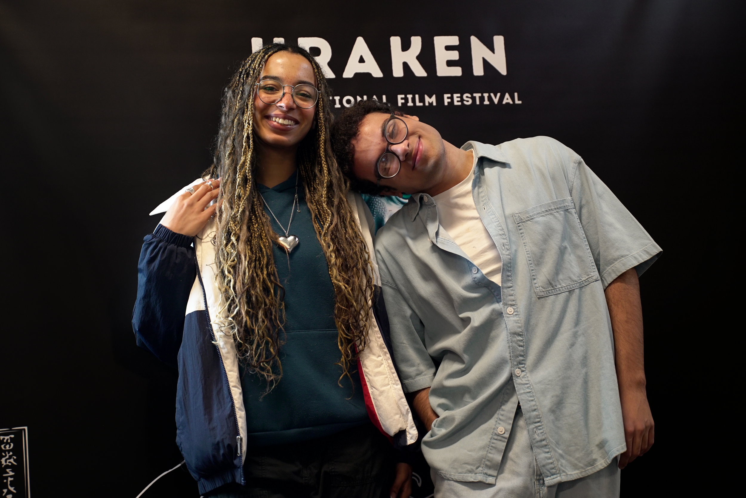 Two people posing at a film festival, one leaning on the other's shoulder, smiling, with a heart-shaped necklace visible.
