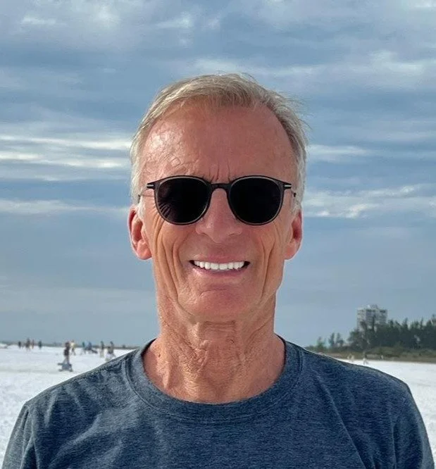 Man wearing sunglasses and a blue shirt, smiling outdoors on a beach with a cloudy sky in the background.