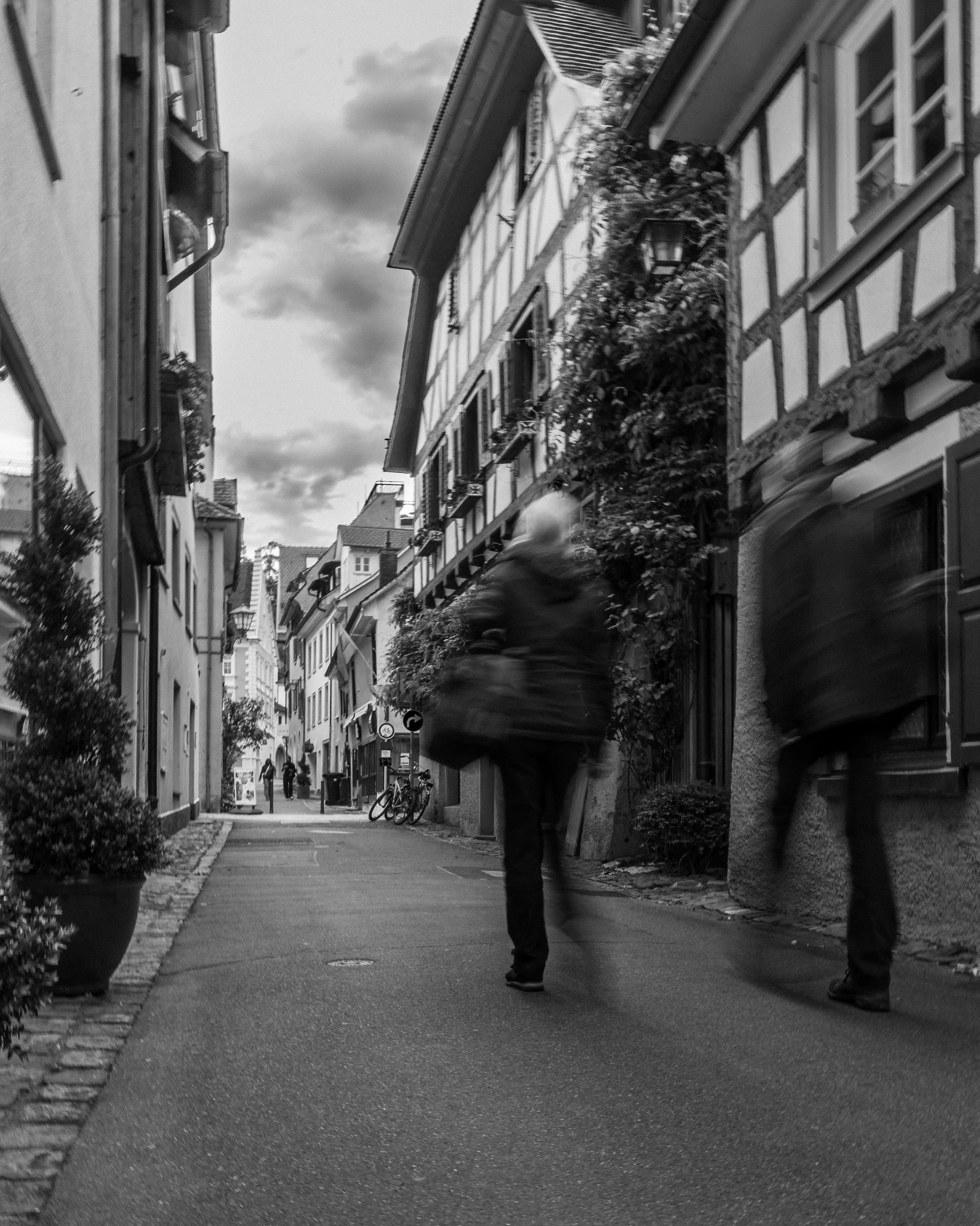 Schwarzweißaufnahme einer engen Altstadtgasse mit Fachwerkhäusern und unscharfen vorbeigehenden Personen.