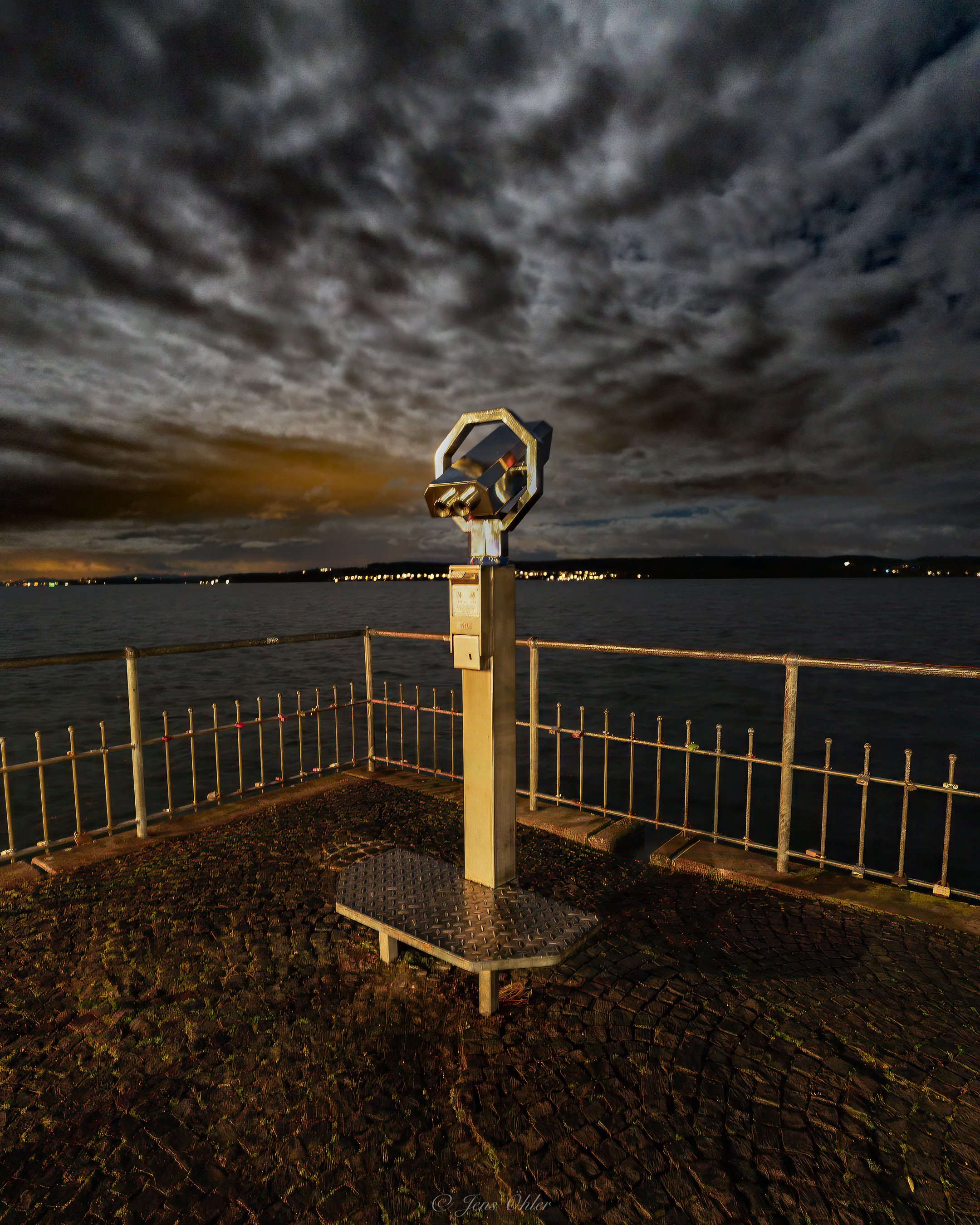 Ein Fernglas auf einem Aussichtspunkt am Wasser bei Nacht, mit bewölktem Himmel und Lichtern im Hintergrund.