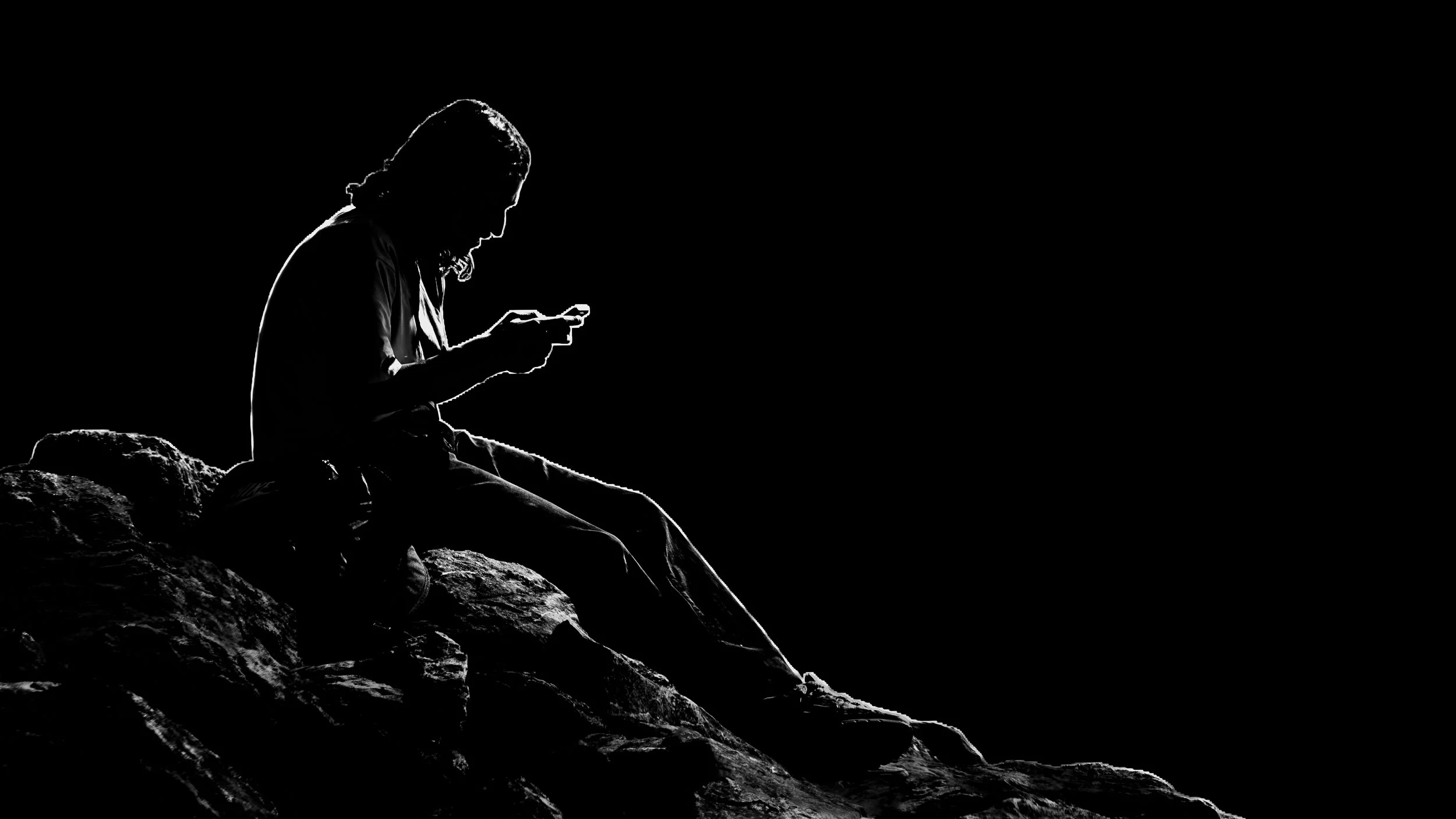 Schwarzweißfotografie einer auf einem Felsen sitzenden Person. Helles Kantenlicht zeichnet ihre Silhouette vor nahezu schwarzem Hintergrund.