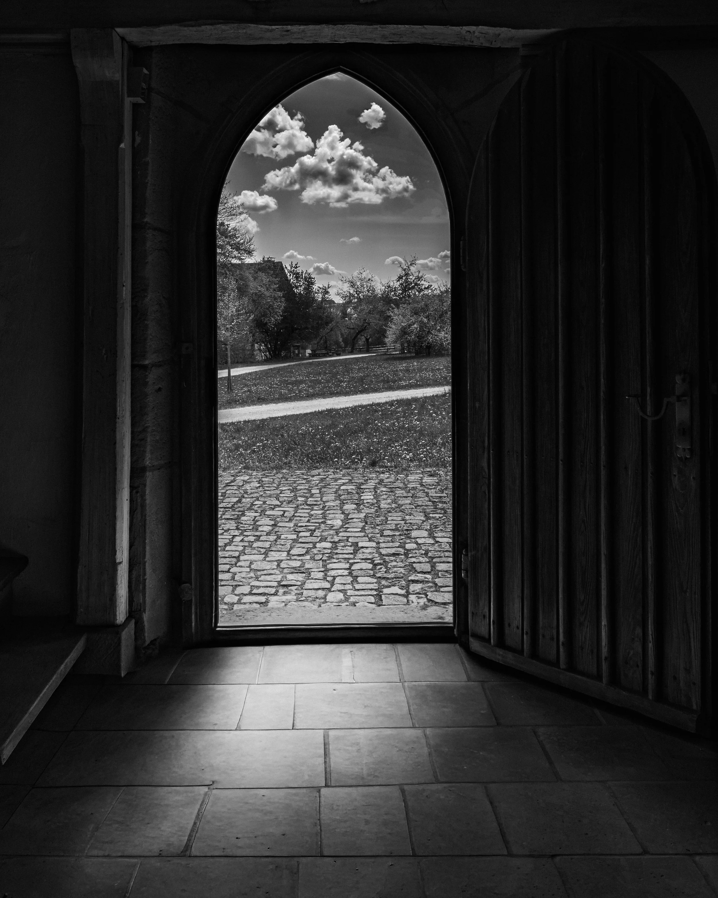 Blick durch ein offenes Holztor auf einen Weg, Bäume und den Himmel mit Wolken. Schwarz-Weiß-Fotografie.