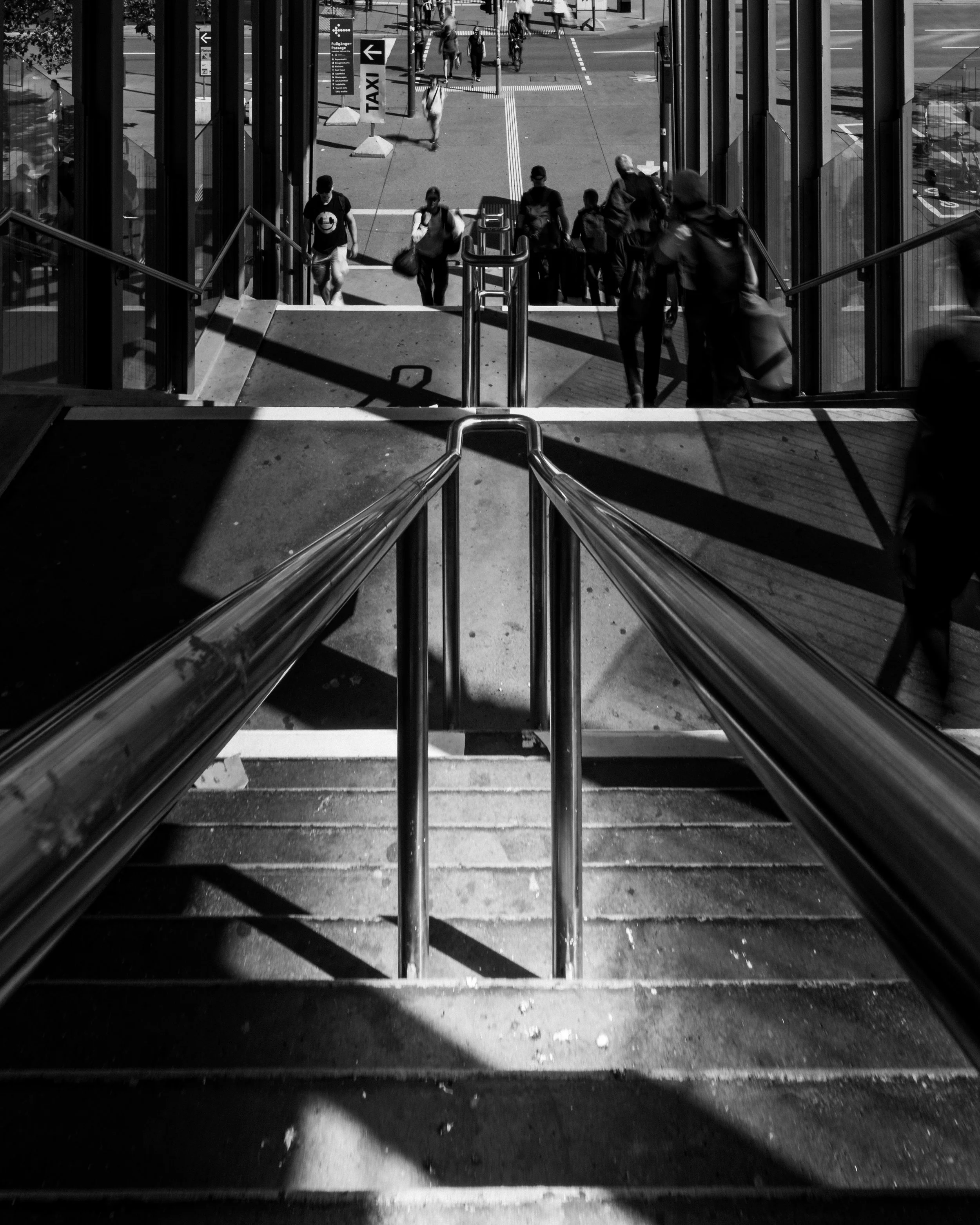 Schwarzweißfoto einer Bahnhofstreppe mit Geländern, starken Schattenlinien und mehreren Menschen im Übergang zwischen Innen- und Außenraum.