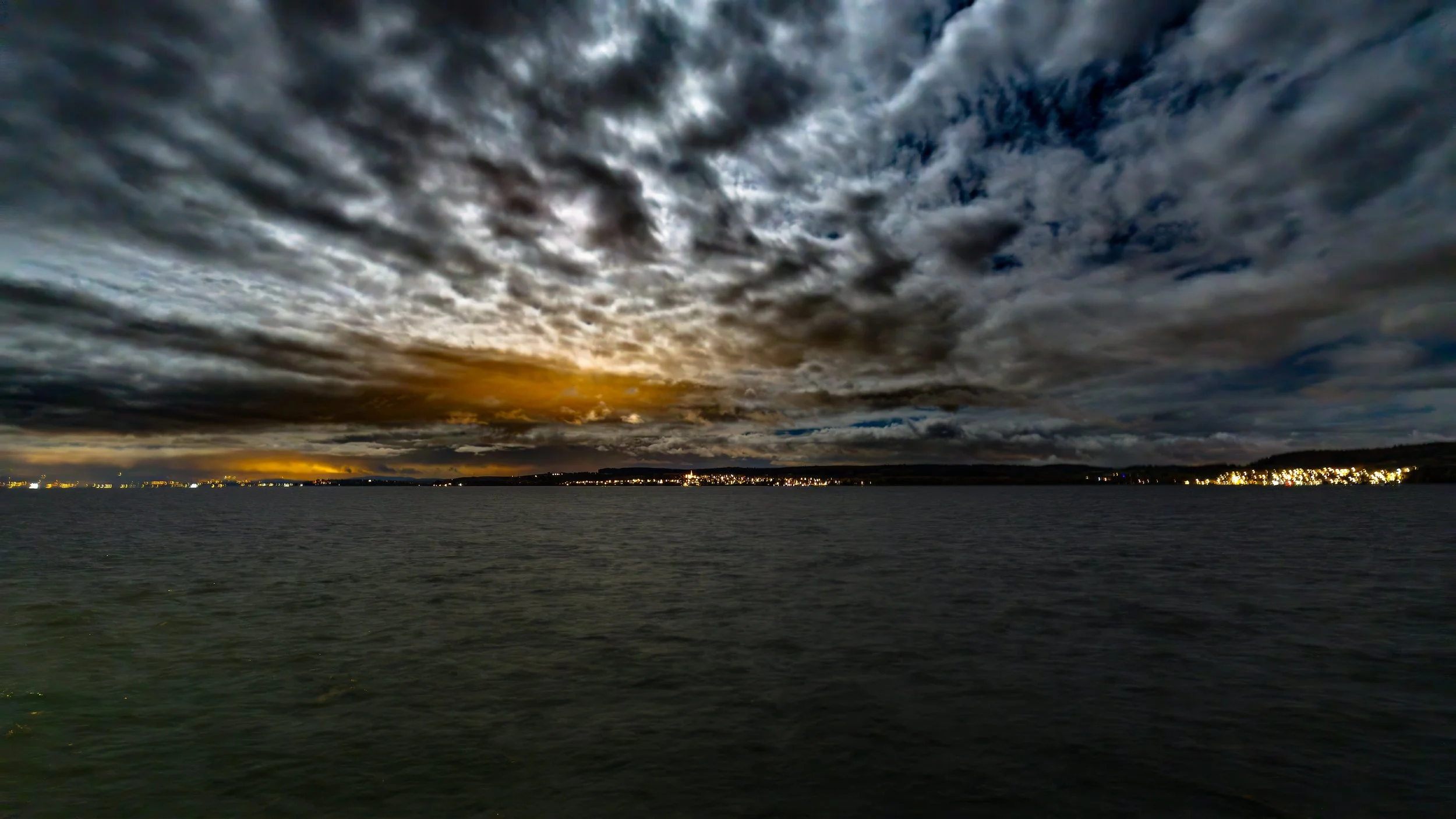 Dunkler Himmel mit dramatischen Wolken über einem ruhigen Gewässer bei Sonnenuntergang, Stadtlichter am Horizont
