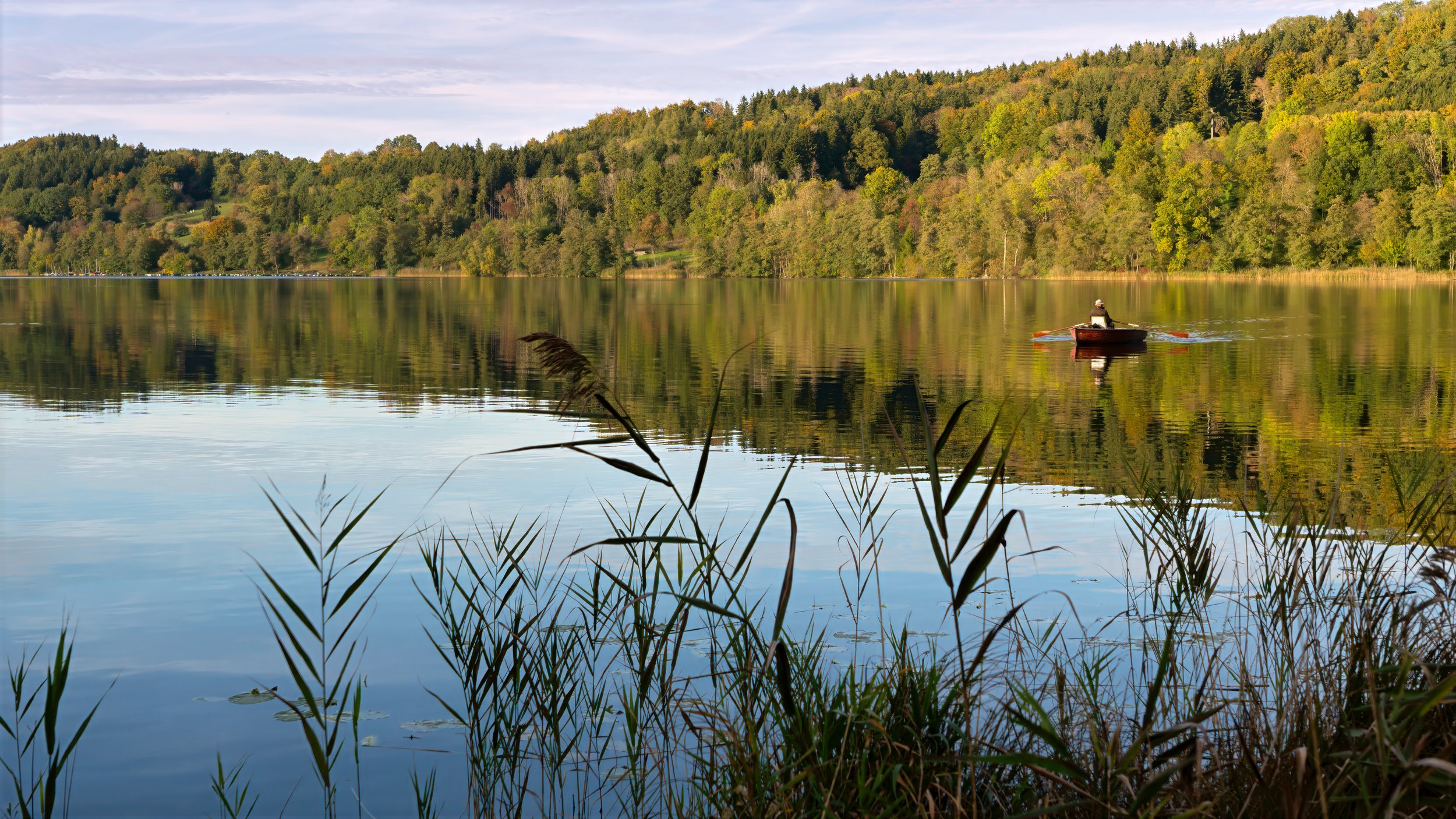 Ein ruhiger See mit einem Ruderboot, umgeben von Bäumen in Herbstfarben, mit reflektiertem Himmel im Wasser.