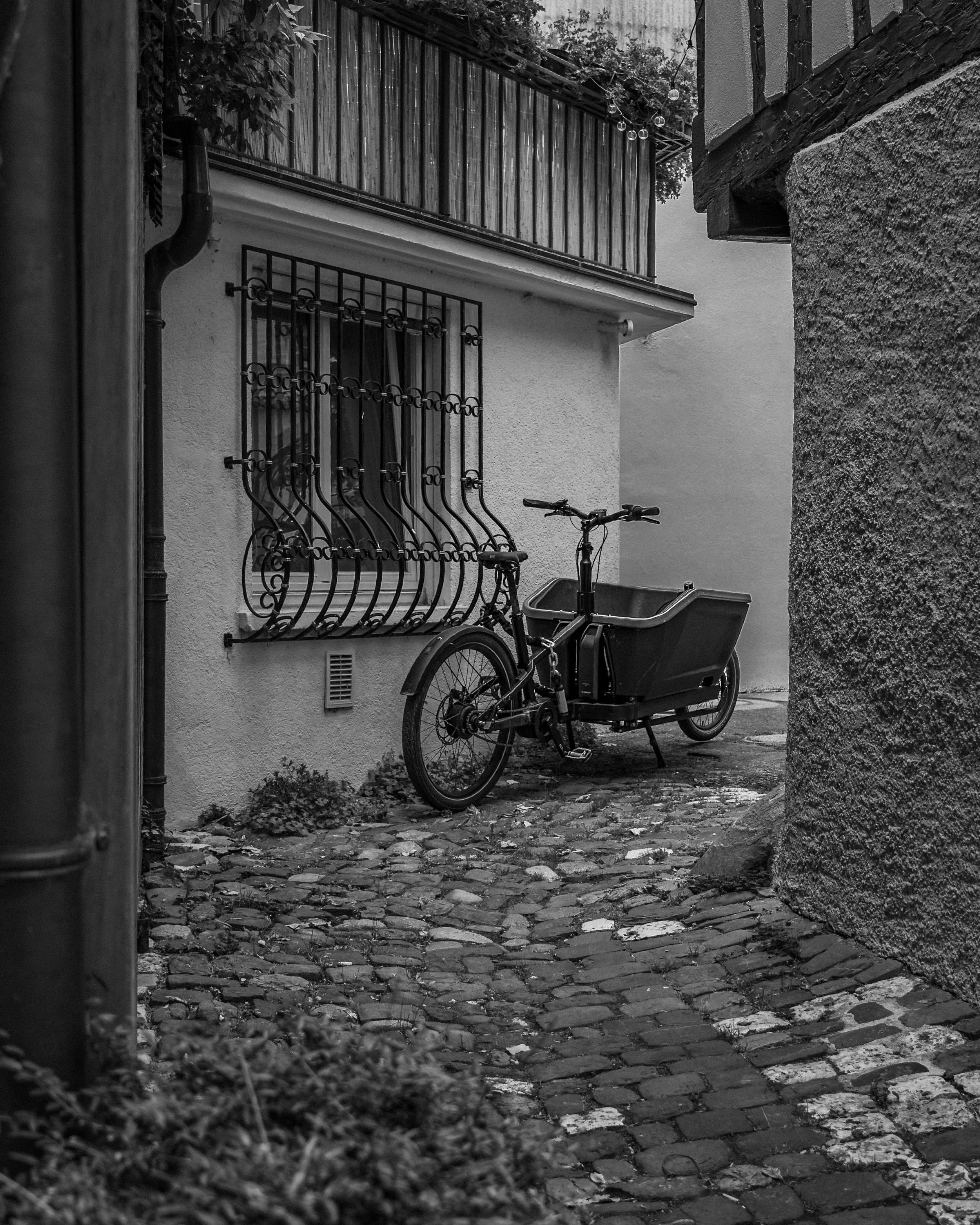Ein Fahrrad mit einem großen Kasten vorne ist geparkt vor einer weißen Hauswand, neben einem Fenster mit dekorativer Gitterverriegelung, im Hintergrund eine Holzterrasse mit Geländern und Pflanzen, im Vordergrund Kopfsteinpflaster auf einem kleinen W