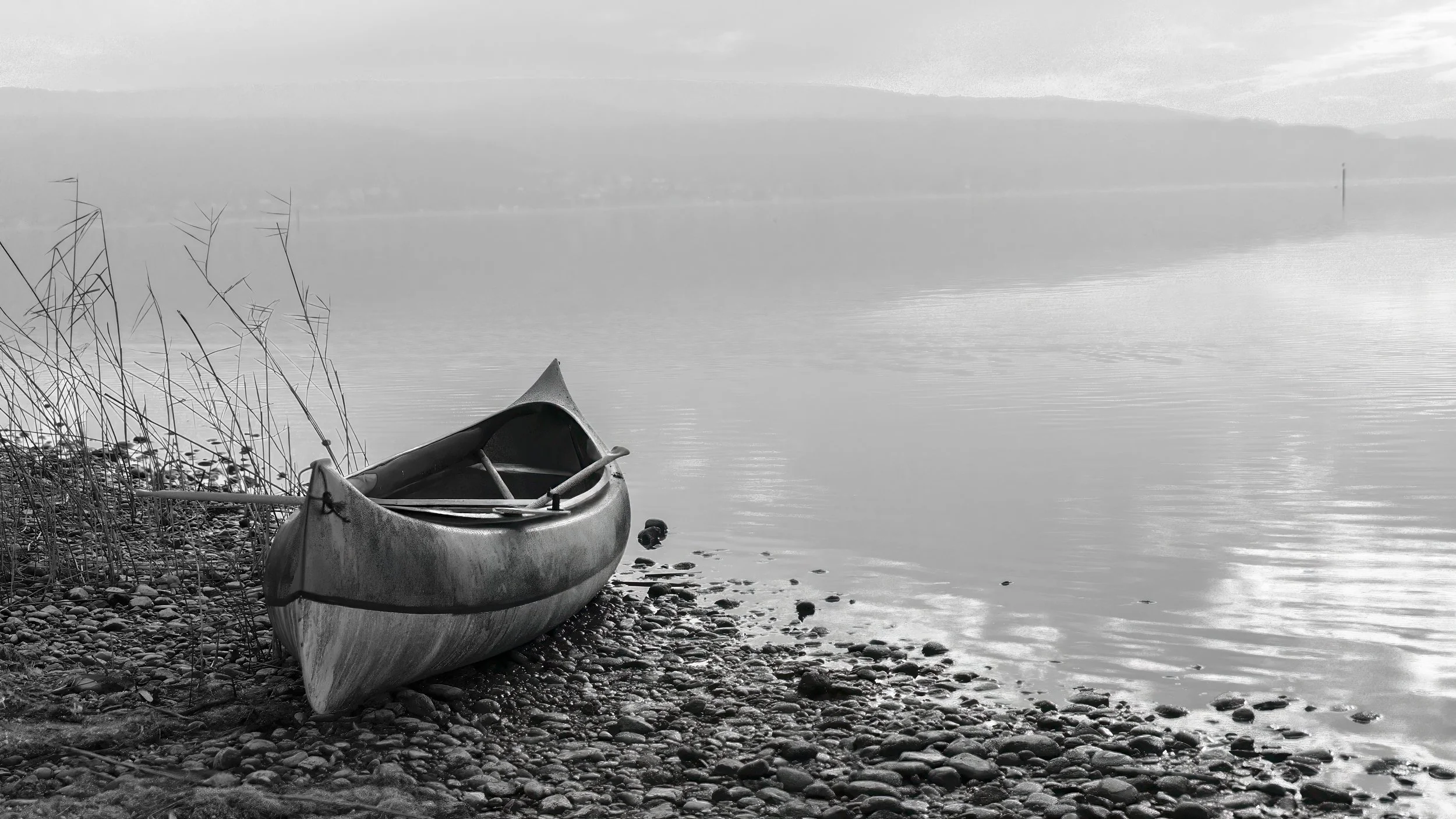 Ein Kanu liegt am Ufer eines ruhigen Sees mit Steinen und wenig Pflanzen, im Hintergrund Berge und eine bewölkte Himmel.