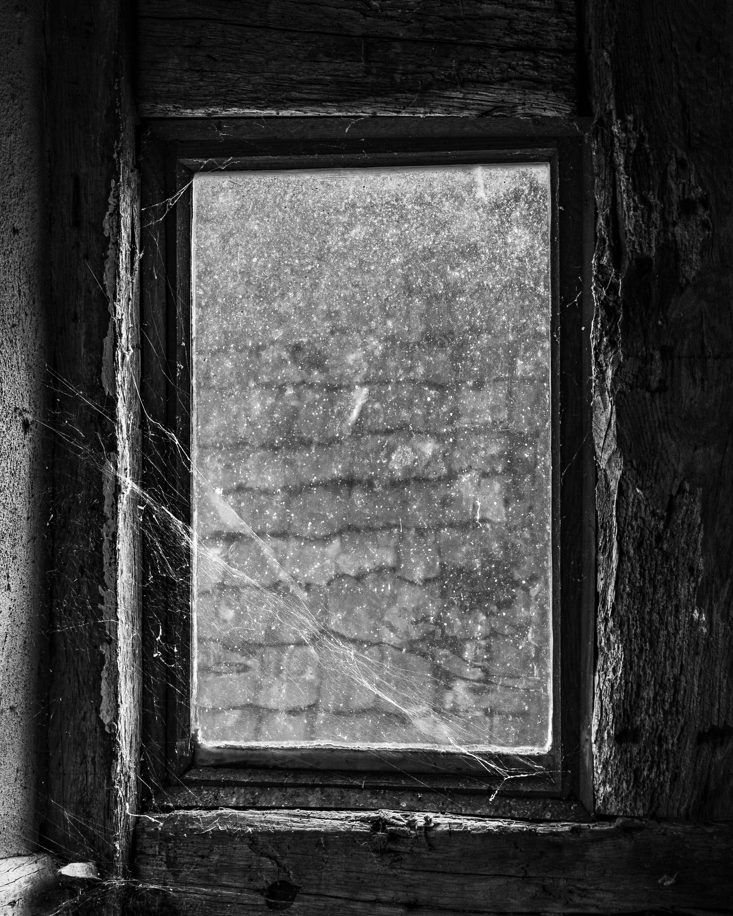 Ein alter, verrotteter hölzerner Rahmen um ein quadratisches Fenster oder eine Öffnung, die mit Staub bedeckt ist. Es gibt Spinnweben an den Ecken und Kanten des Rahmens, und die Struktur sieht verfallen und vernachlässigt aus.