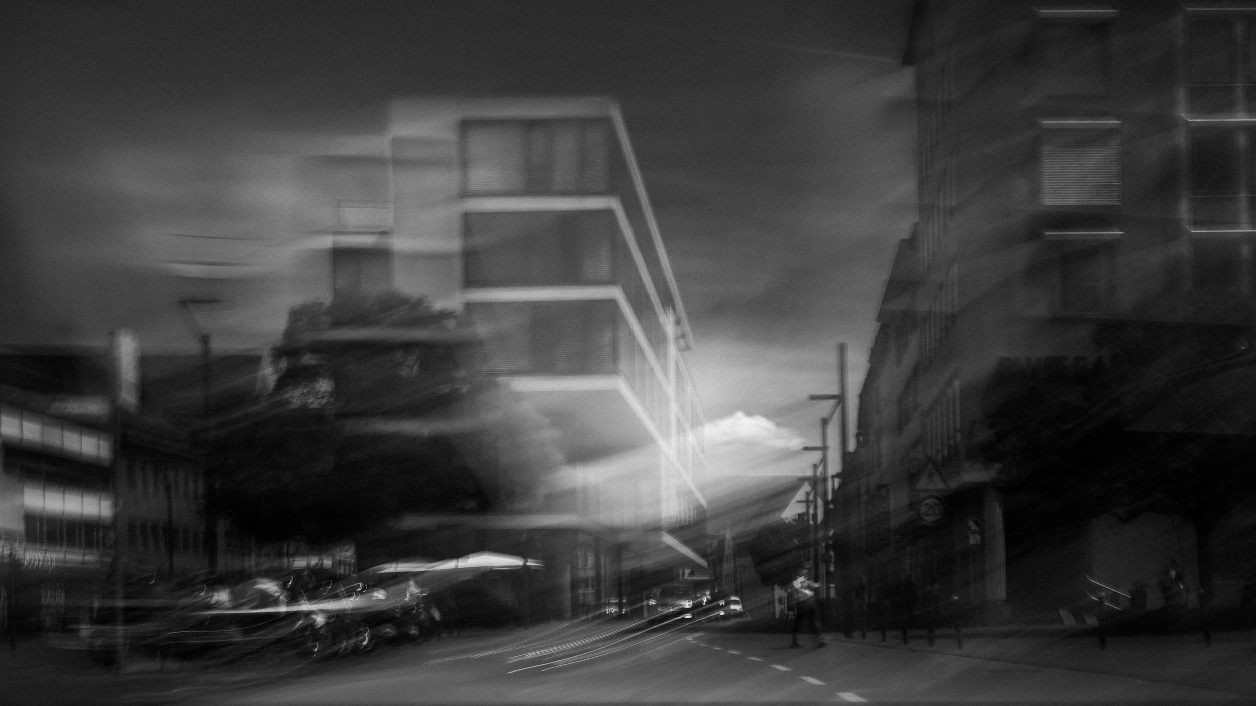Schwarz-weiß-Foto einer urbanen Straße mit modernen Gebäuden, Autos und Stromleitungen bei Nacht, mit Lichtspuren durch langsame Verschlusszeit.