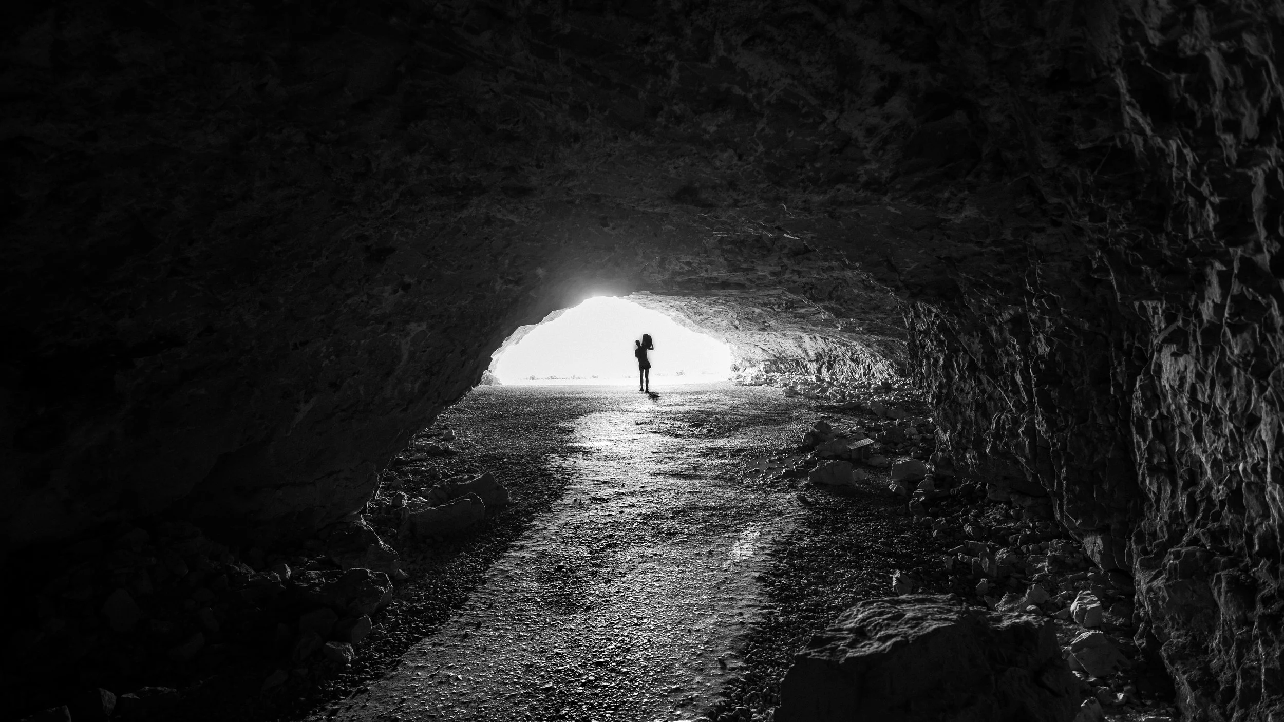Schwarzweißfoto einer Höhle mit hellem Ausgang, rauer Felsstruktur und einer kleinen Silhouette im Gegenlicht.