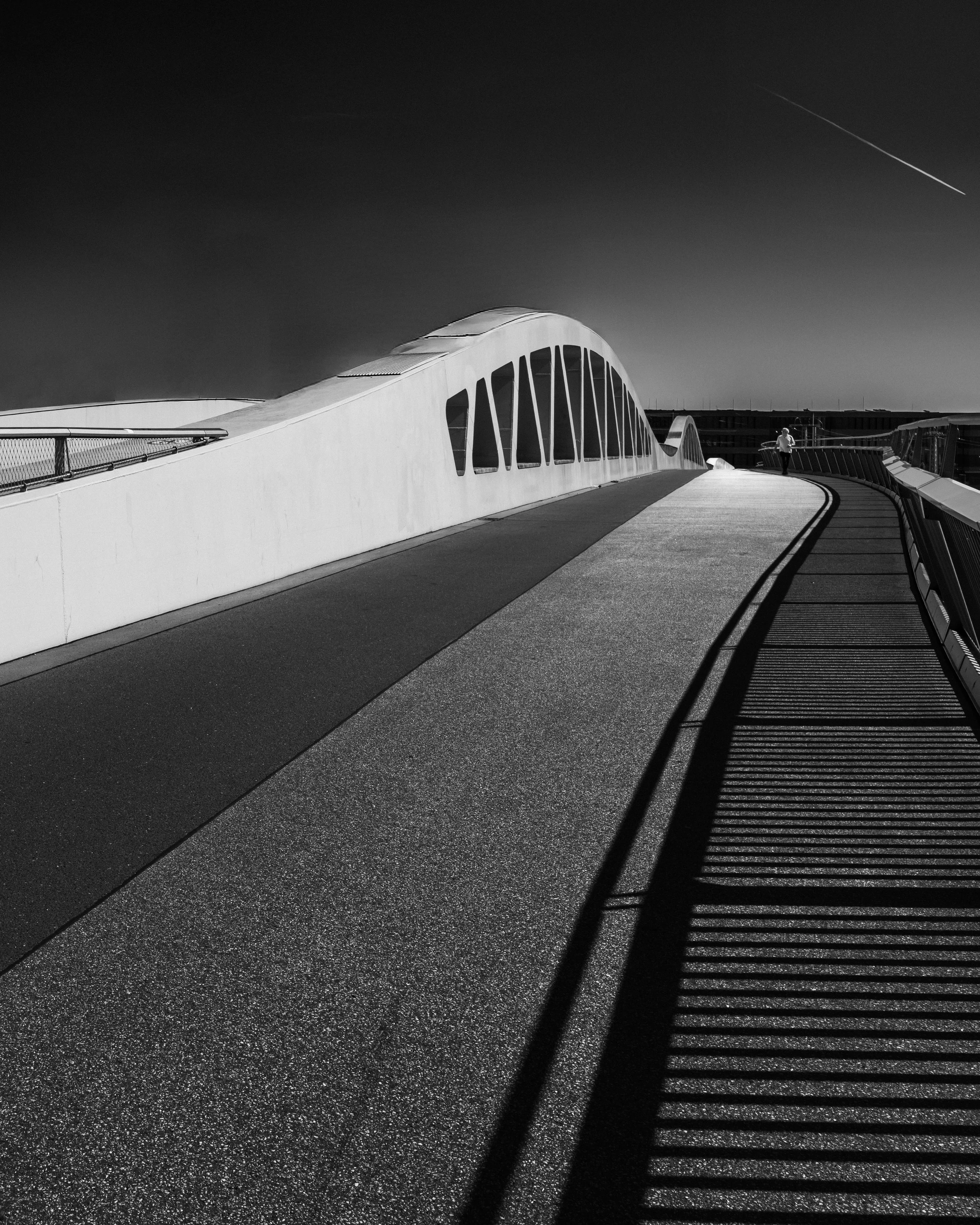 Schwarzweißaufnahme einer modernen Fußgängerbrücke mit geschwungener Linie, starkem Schattenmuster und weiter Blickrichtung.