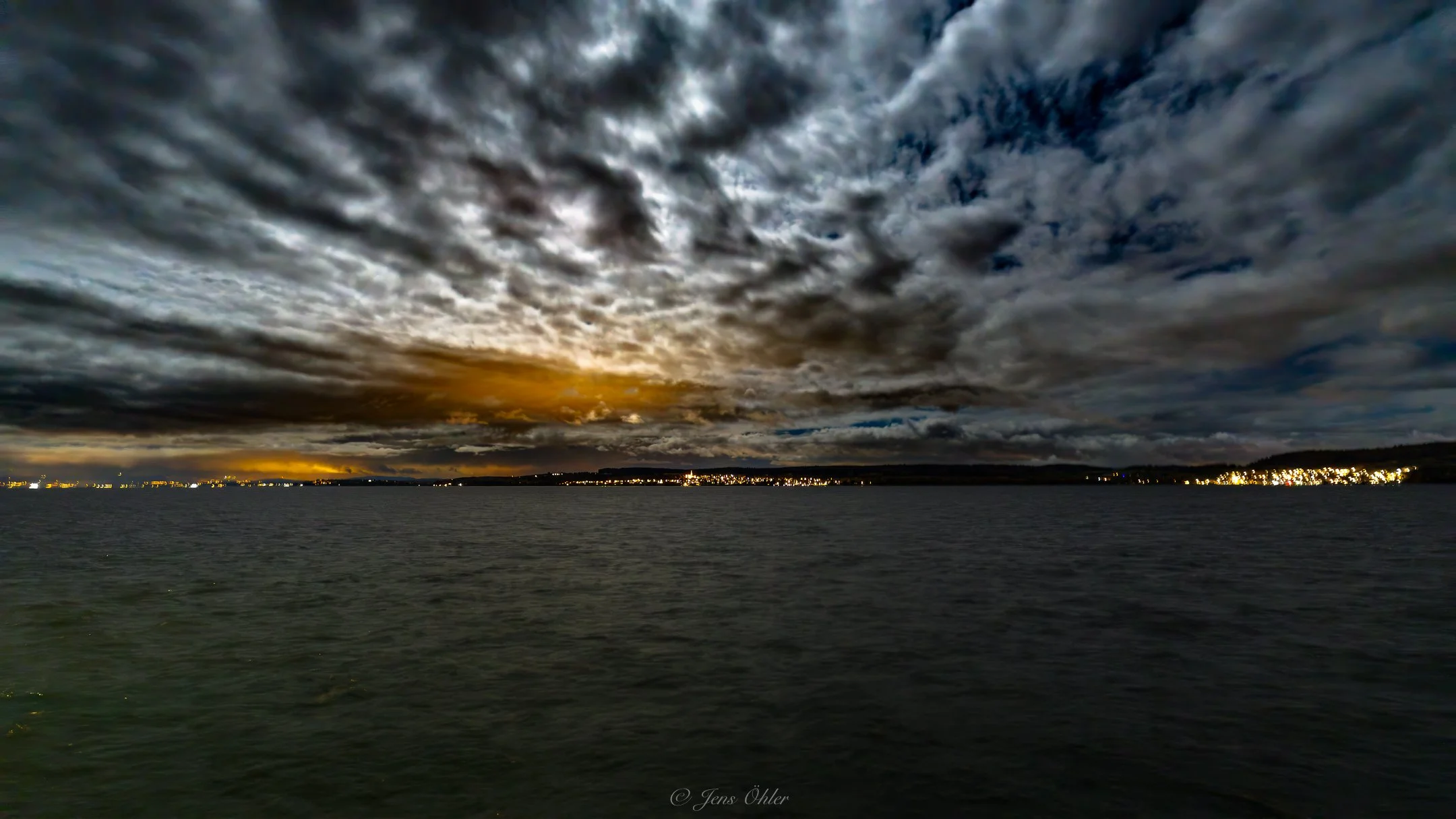 Dunkle Wolken über einem ruhigen See bei Sonnenuntergang, mit Lichtern entlang des Horizonts.