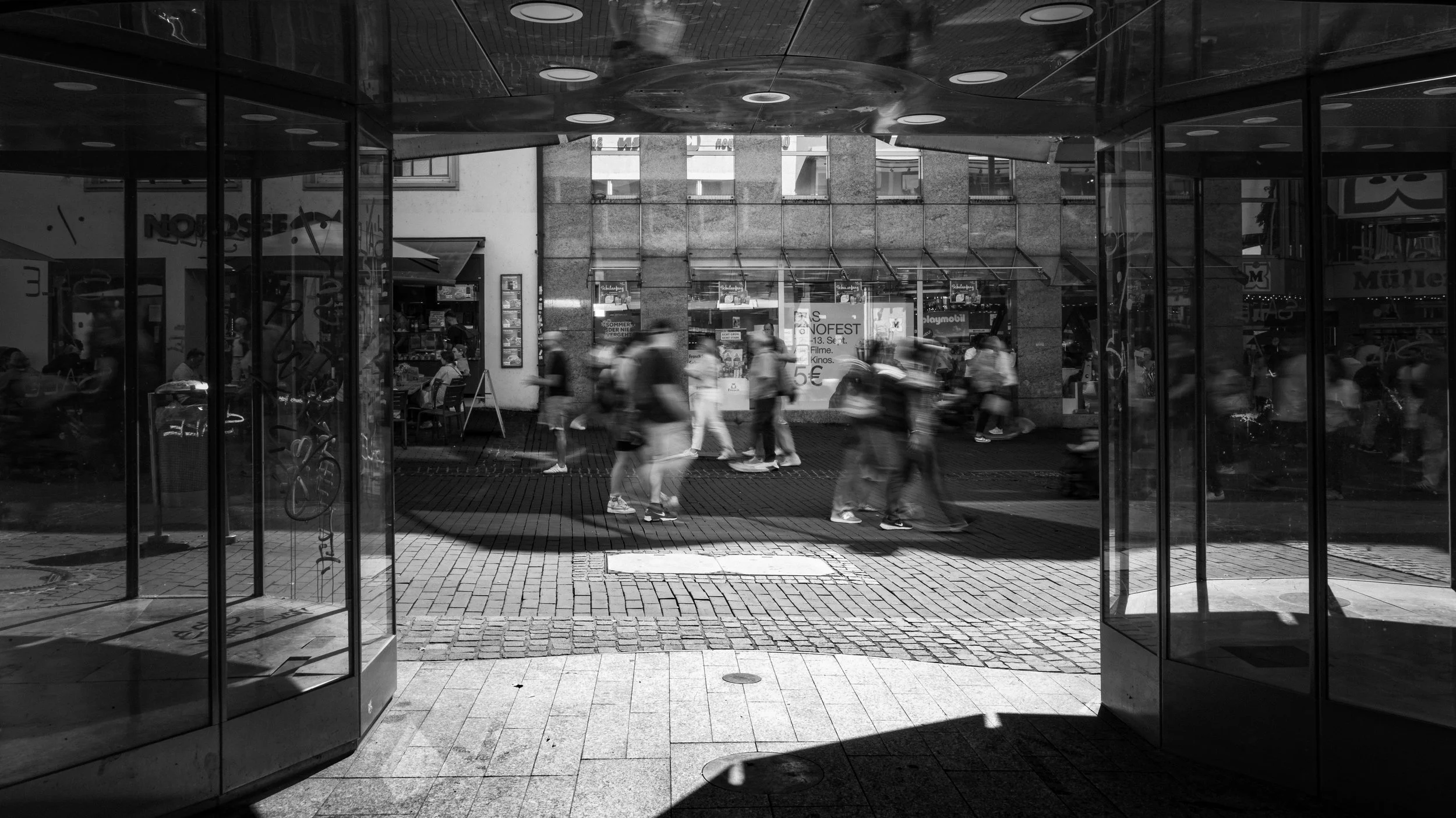 Blick durch eine Glastür auf eine belebte Fußgängerzone mit mehreren Menschen, die vor einem Geschäft vorbeigehen, im Hintergrund ein Gebäude mit Schaufenstern und Werbetafeln.
