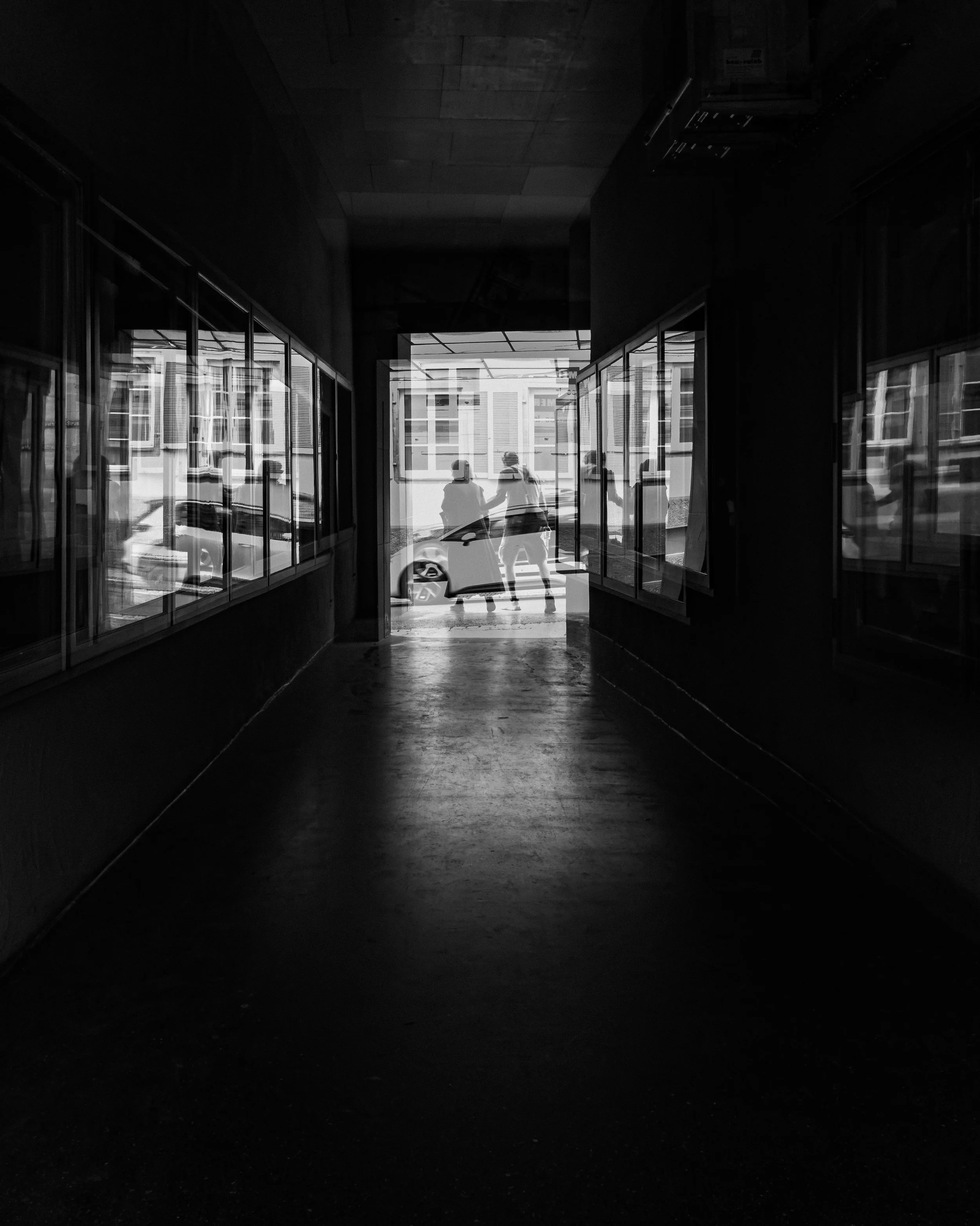 Schwarzweißaufnahme eines dunklen Durchgangs mit Spiegelungen, tiefen Schatten und hellen silhouettenhaften Figuren am Ausgang.