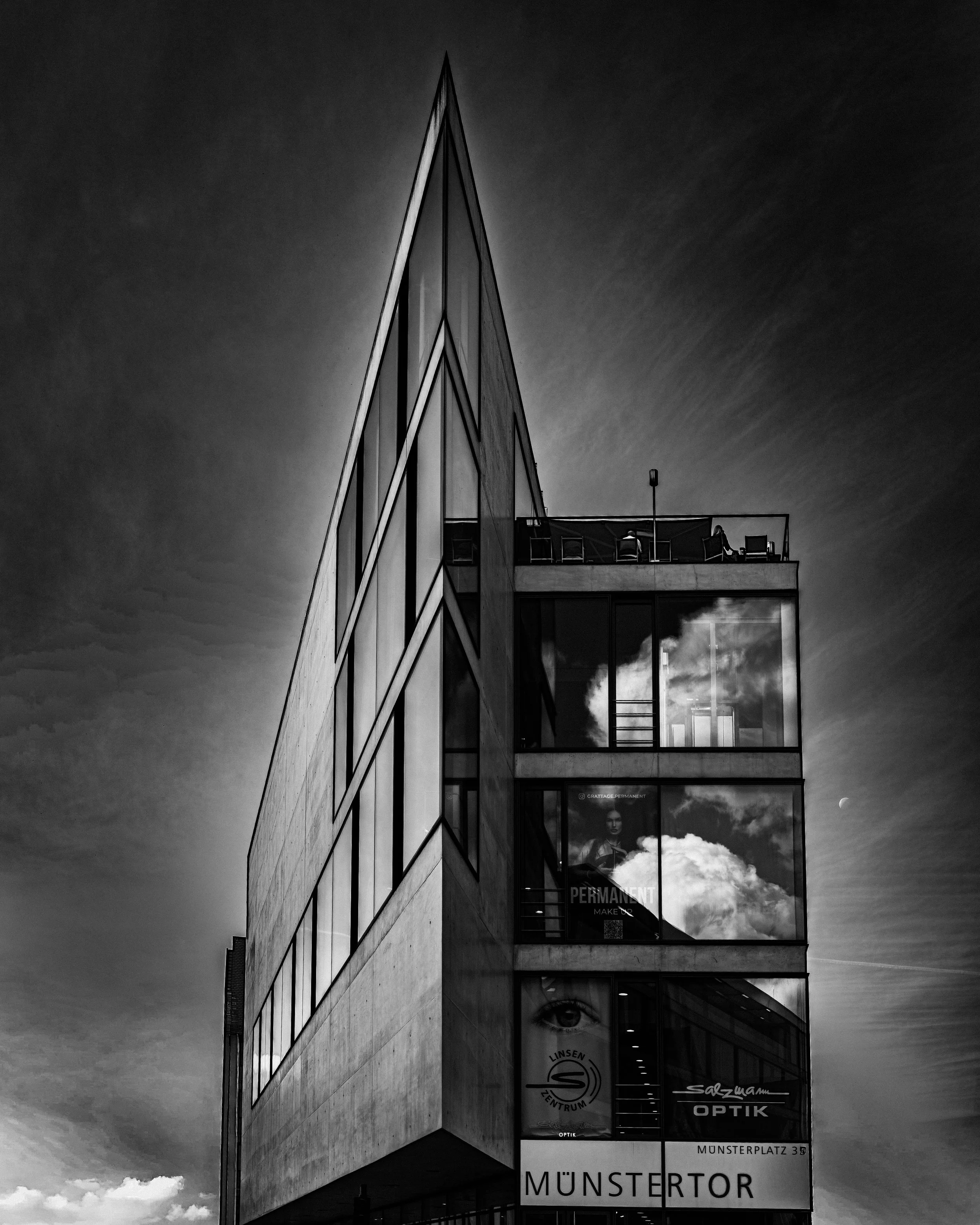 Schwarzweißaufnahme des Münstertors in Ulm mit spitz zulaufender Architektur, Glasfassade, Wolkenreflexionen und kleinem Mond am Himmel.