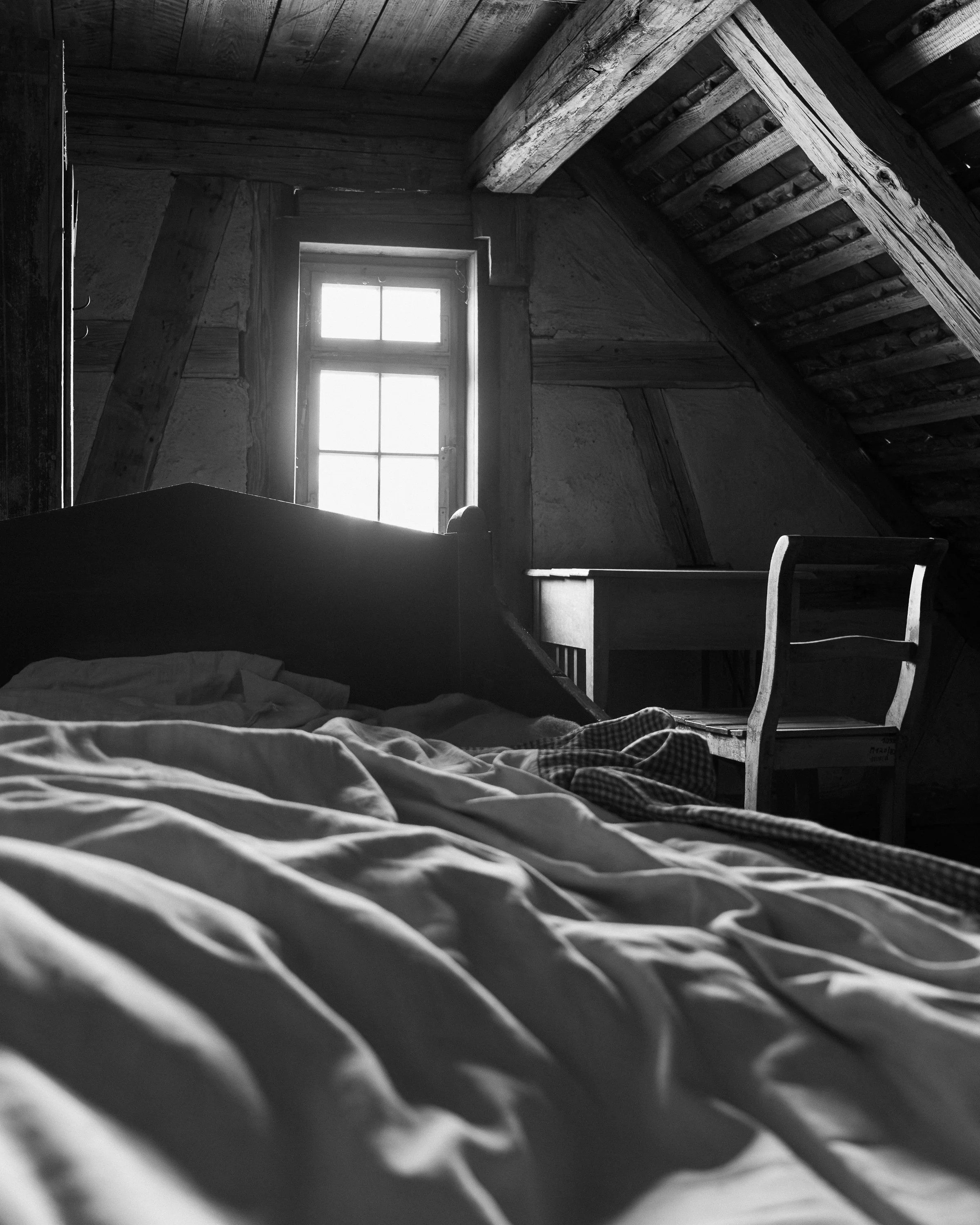 Schwarzweißfotografie eines historischen Schlafraums. Im Vordergrund ein ungemachtes Bett, dahinter ein helles Fenster und ein Holzstuhl unter den schrägen Dachbalken.
