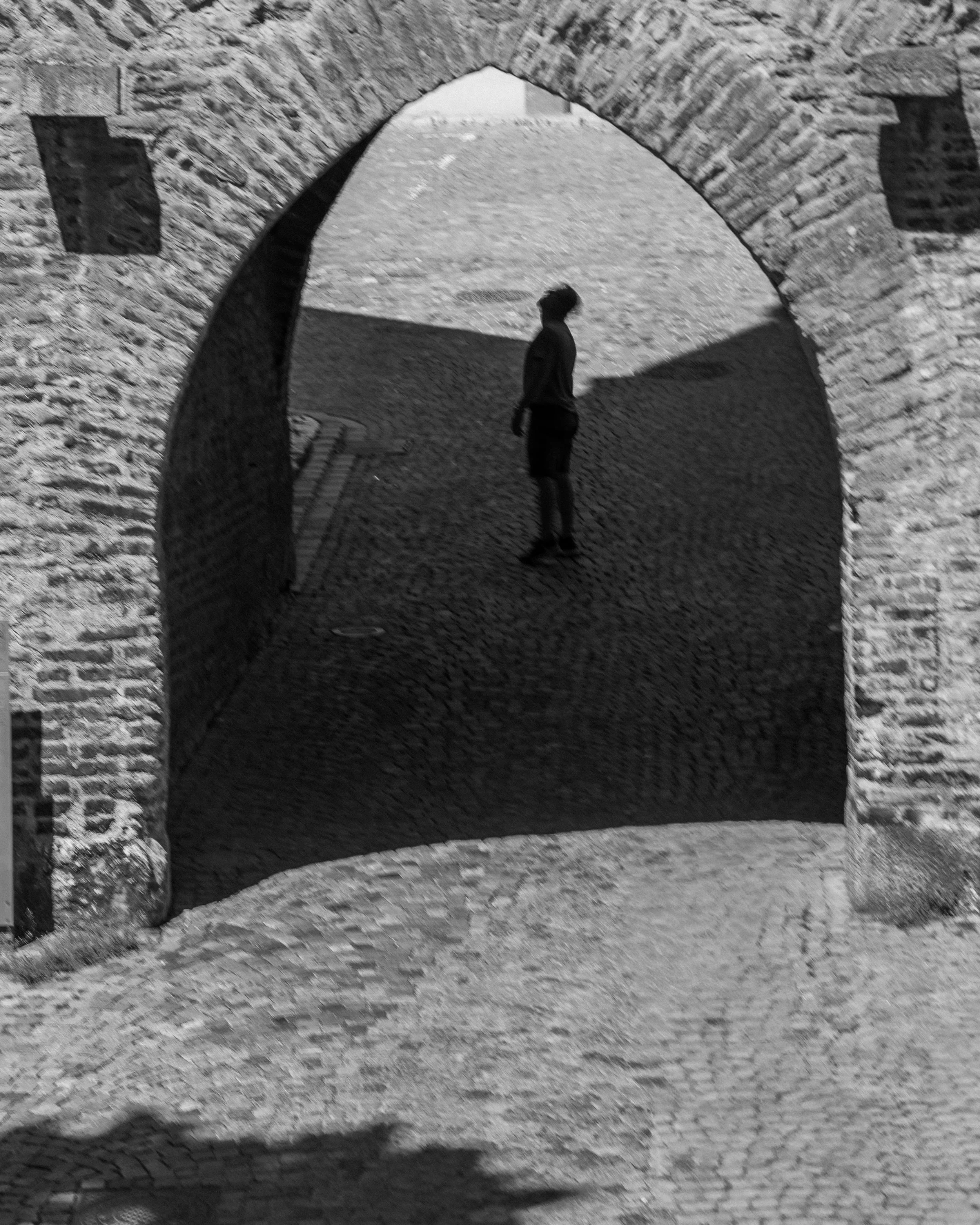 Schwarzes und weißes Foto eines Menschen, der unter einer Steinbrücke steht. Der Mensch schaut nach oben, während die Brücke aus Steinen besteht und einen Bogen bildet. Das Bild ist in Schwarz-Weiß gehalten.