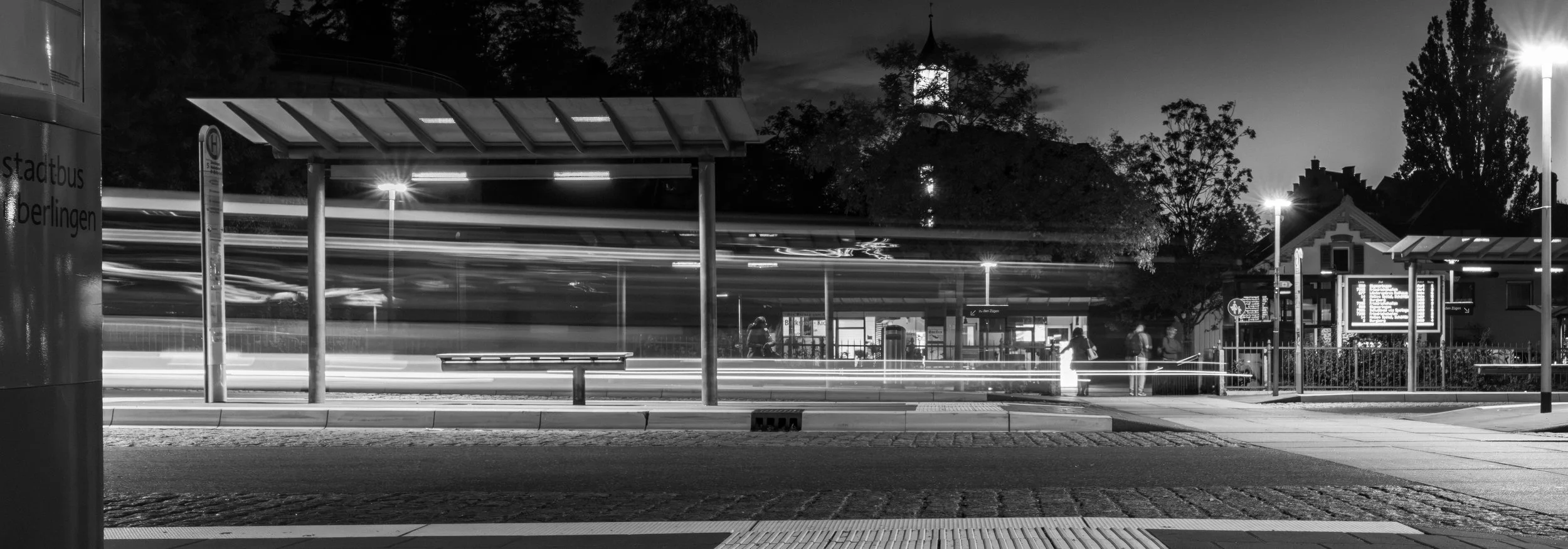 Schwarz-Weiß-Foto eines Bushalts in einer Stadt bei Nacht, mit Lichtspuren von vorbeifahrenden Fahrzeugen, wartenden Menschen und übersichtlichem Anzeigetafel.