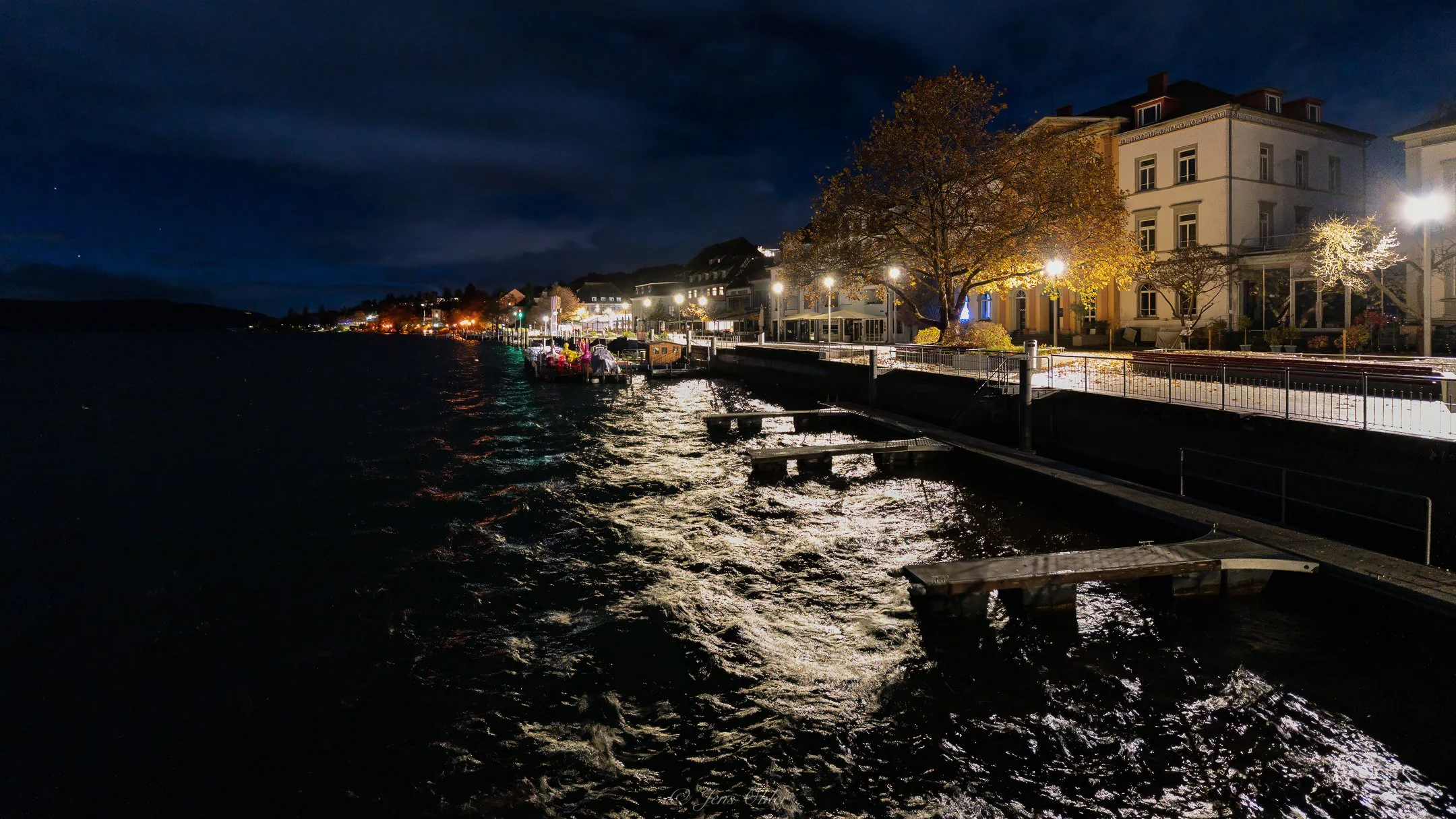 Nachtbild eines beleuchteten Ufers mit Gebäuden, Bäumen und Wasserseiten, reflektierenden Lichtern auf dem Wasser, dunklem Himmel und Wasser, ruhigem Wasser und belebtem Ufer.