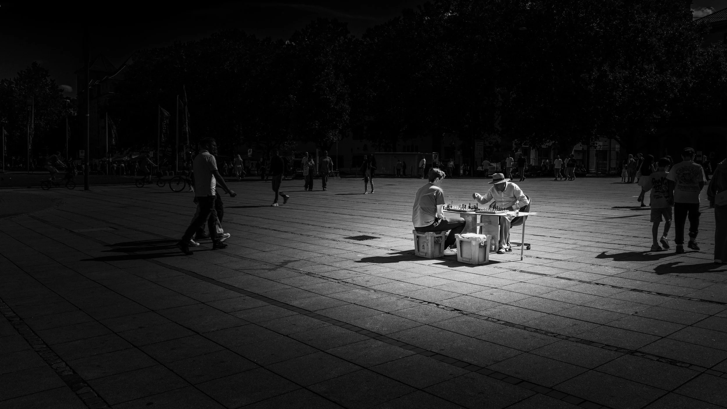 Schwarzweißfoto zweier Schachspieler auf einem großen Platz, umgeben von Licht, Schatten und vorbeigehenden Menschen.