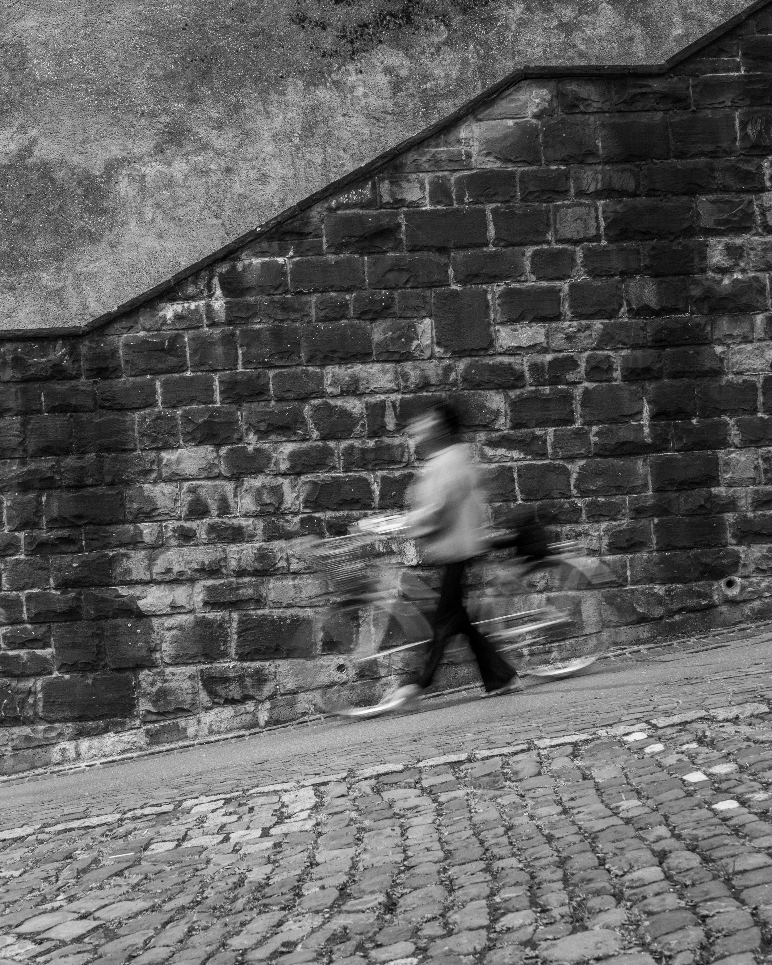 Schwarzweißfoto einer bewegten Person mit Fahrrad vor einer alten Steinmauer und Kopfsteinpflaster.