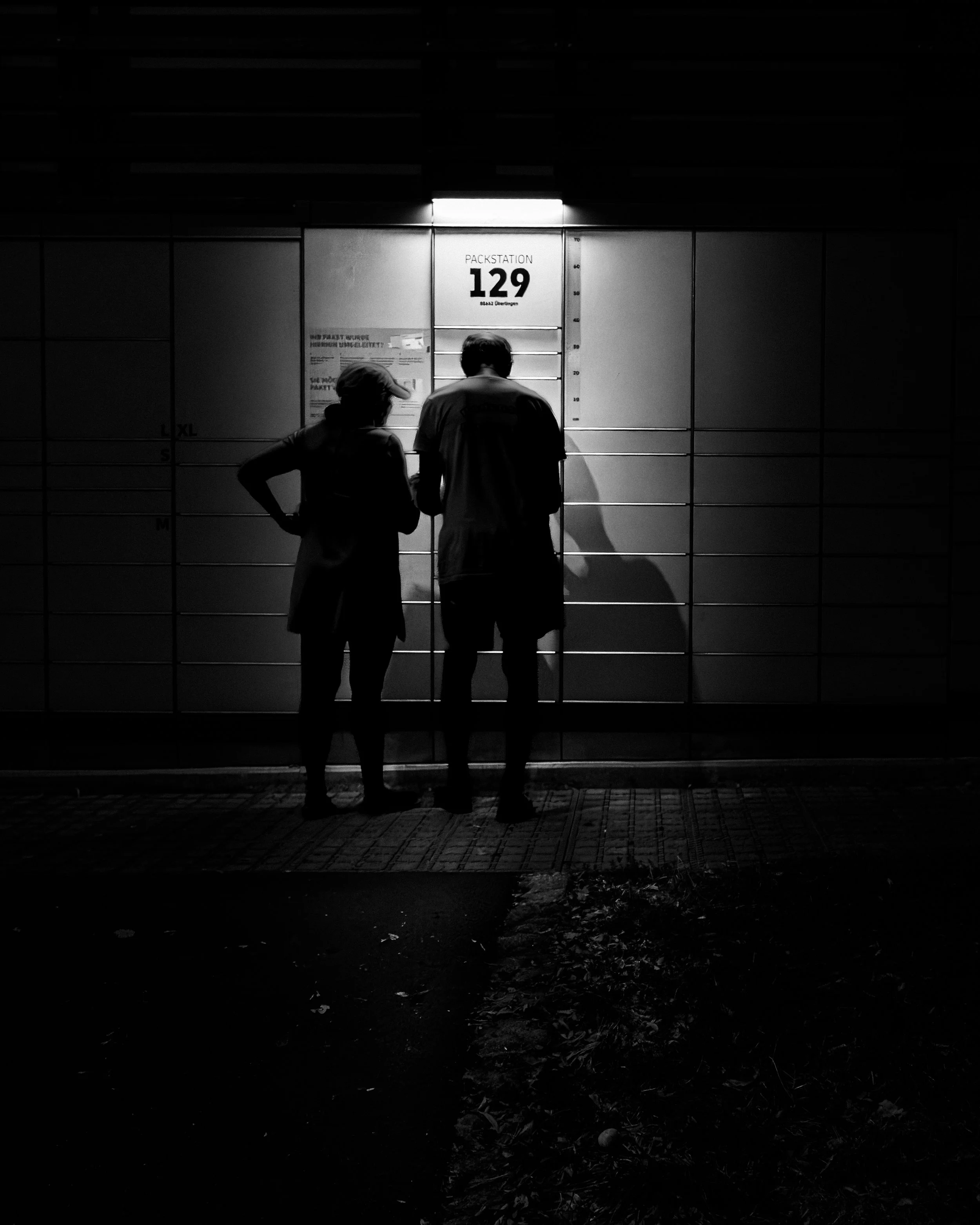 Zwei Personen stehen vor einer Packstation mit dunklem Hintergrund. Die Packstation ist beleuchtet und trägt die Beschriftung „Packstation 129“. Die Szene ist in Schwarz-Weiß gehalten.