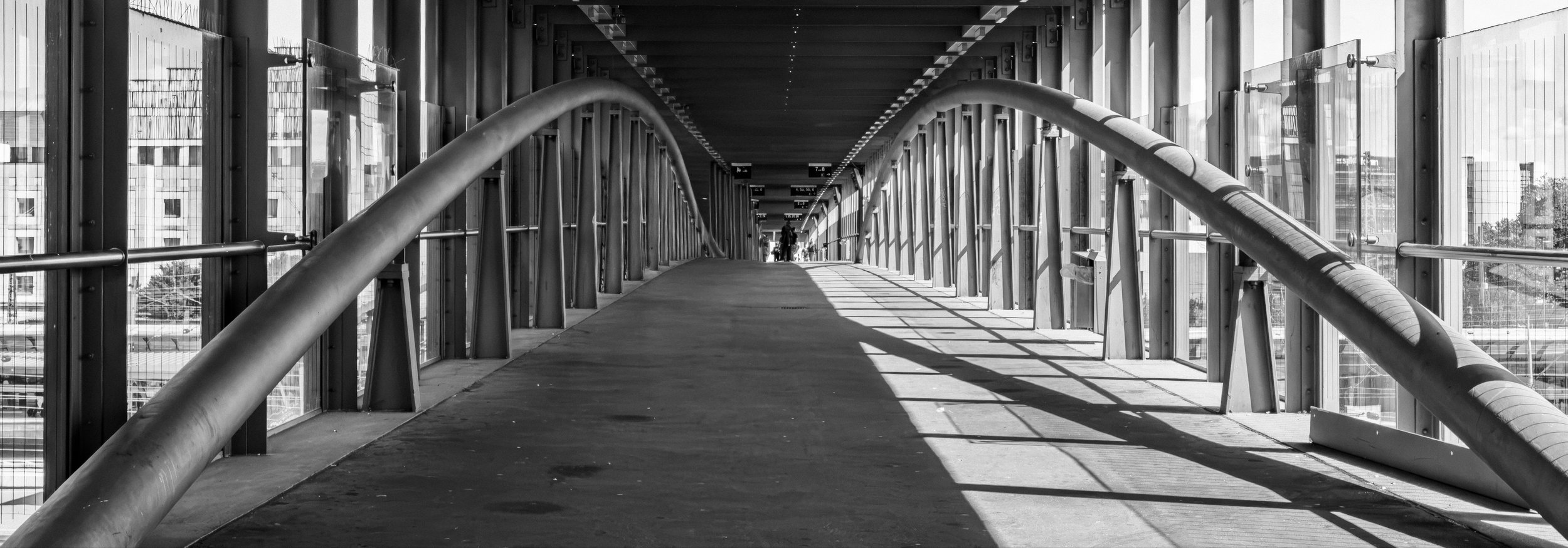 Blick durch einen modernen, geschwungenen Überweg mit großen Glaswänden, Schatten und Silhouetten von Personen am Ende