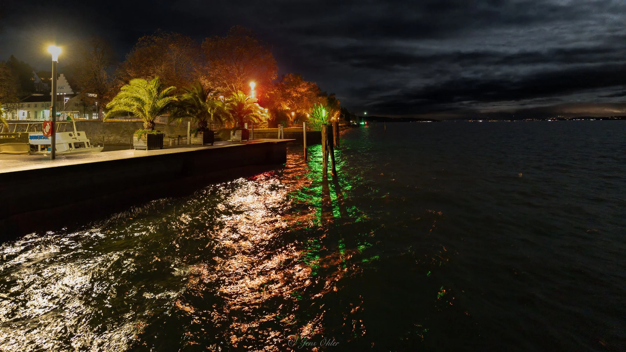 Nachtaufnahme eines Hafens mit beleuchteten Bäumen, Lampen und einem Wasserweg, reflektierende Lichter auf dem Wasser, dunkler Himmel.