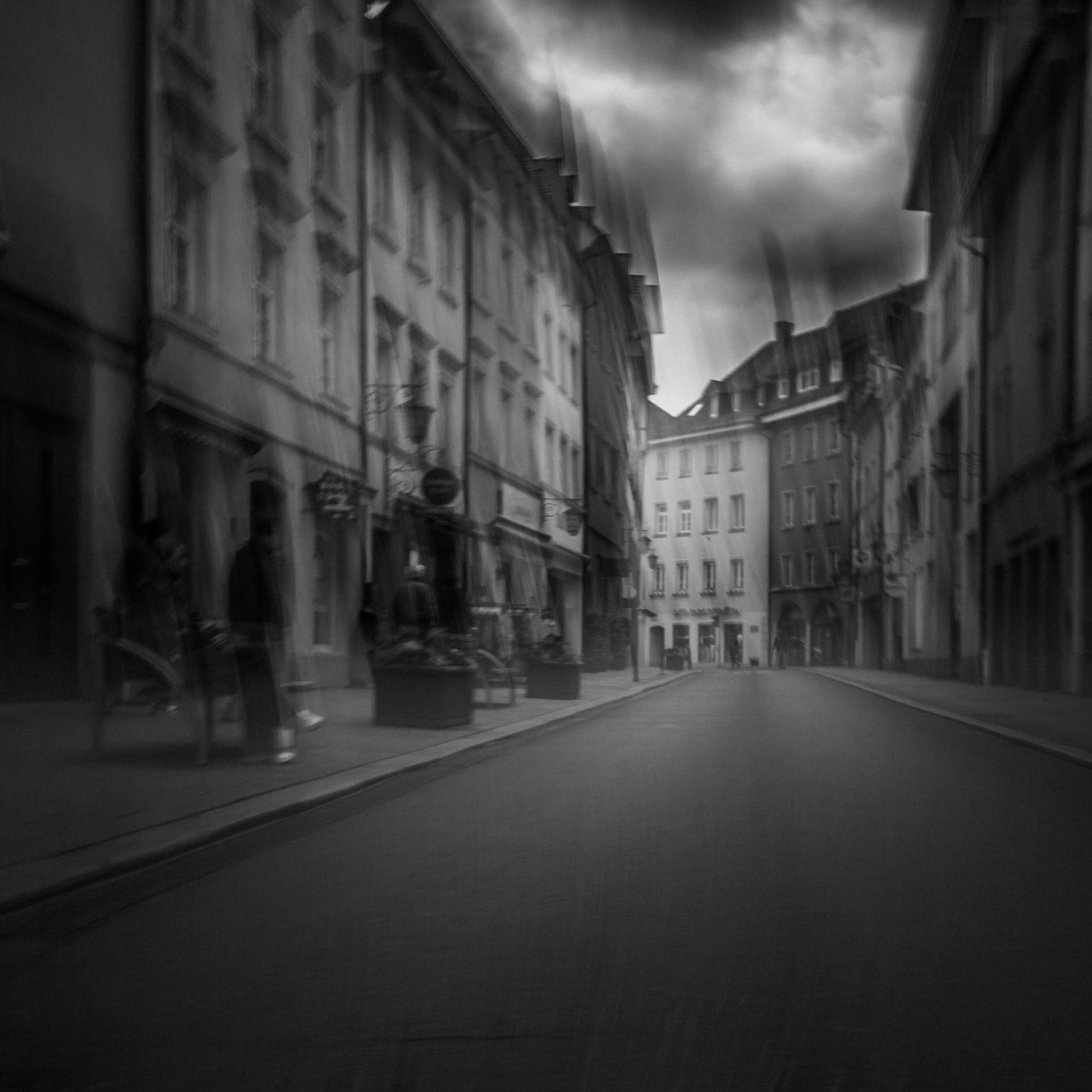 Schwarz-weiß-Bild einer Stadtstraße mit alten Gebäuden, Himmel und Wolken im Hintergrund, keine Fahrzeuge sichtbar.