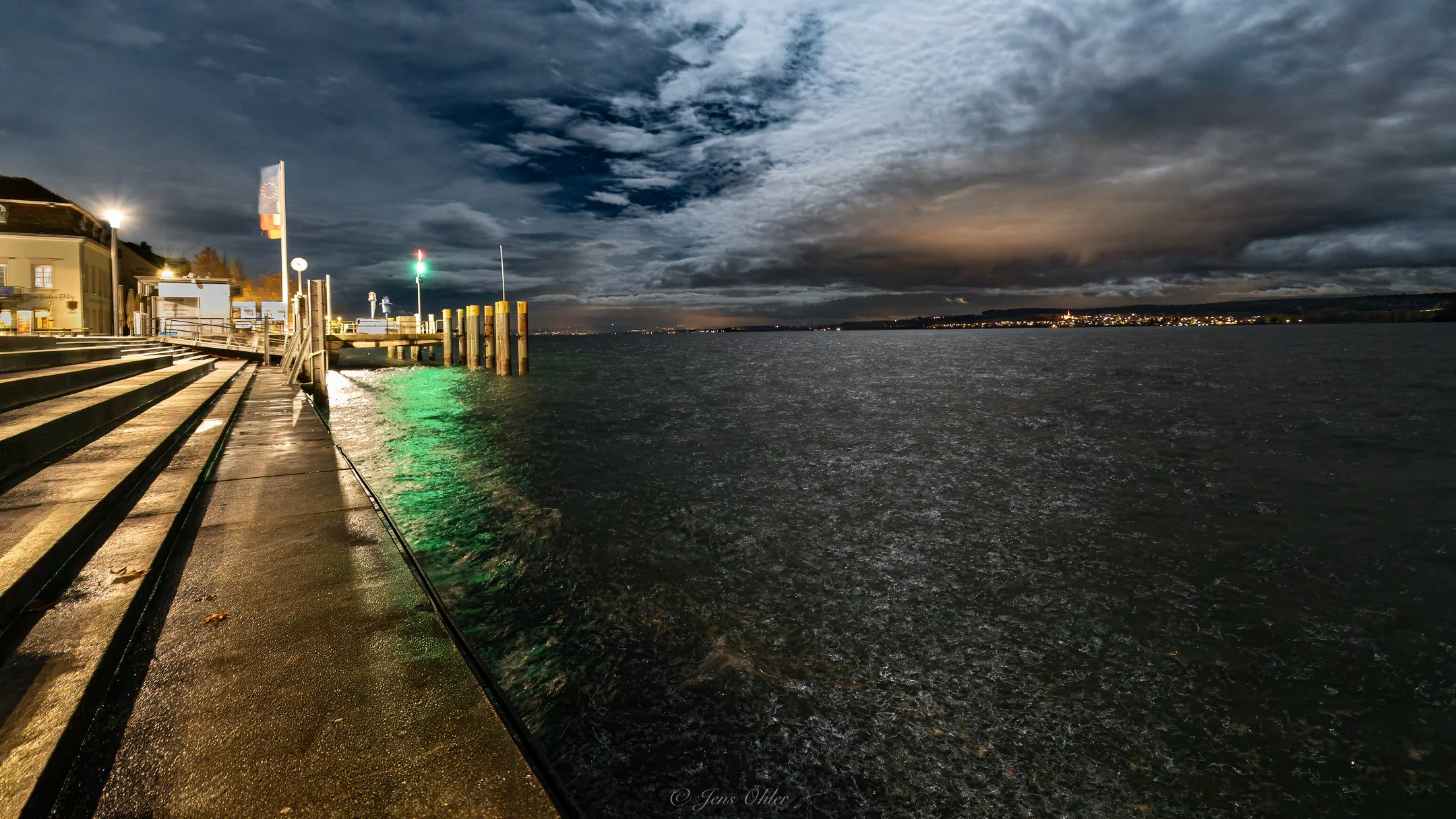 Abendansicht eines Hafens mit einer Pontonbrücke, beleuchtete Gebäude und dunkle Wolken am Himmel, reflektierende Wasserfläche