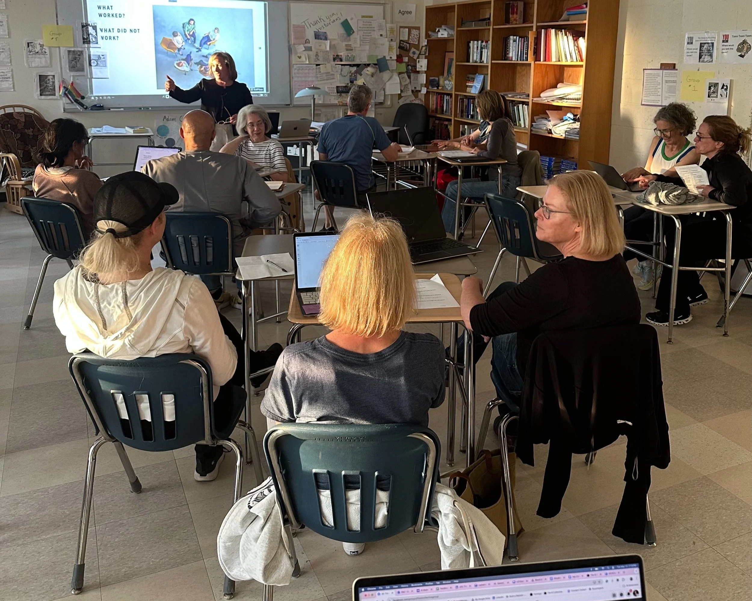 A classroom with adult students seated at tables, listening to a female instructor standing in front of a whiteboard and projector screen.