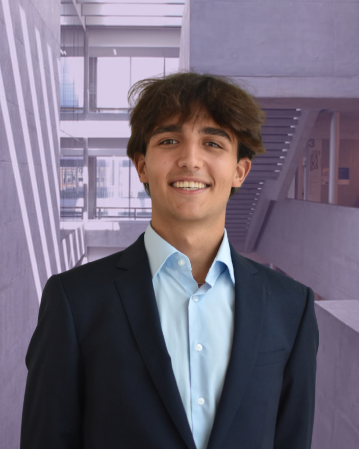 Smiling person with curly hair and patterned shirt standing against a plain background.