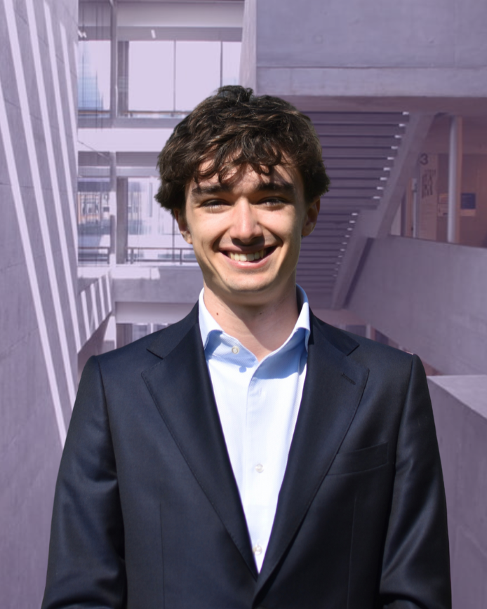 Person in a suit standing outdoors in front of a building with a textured facade and grass in the foreground.