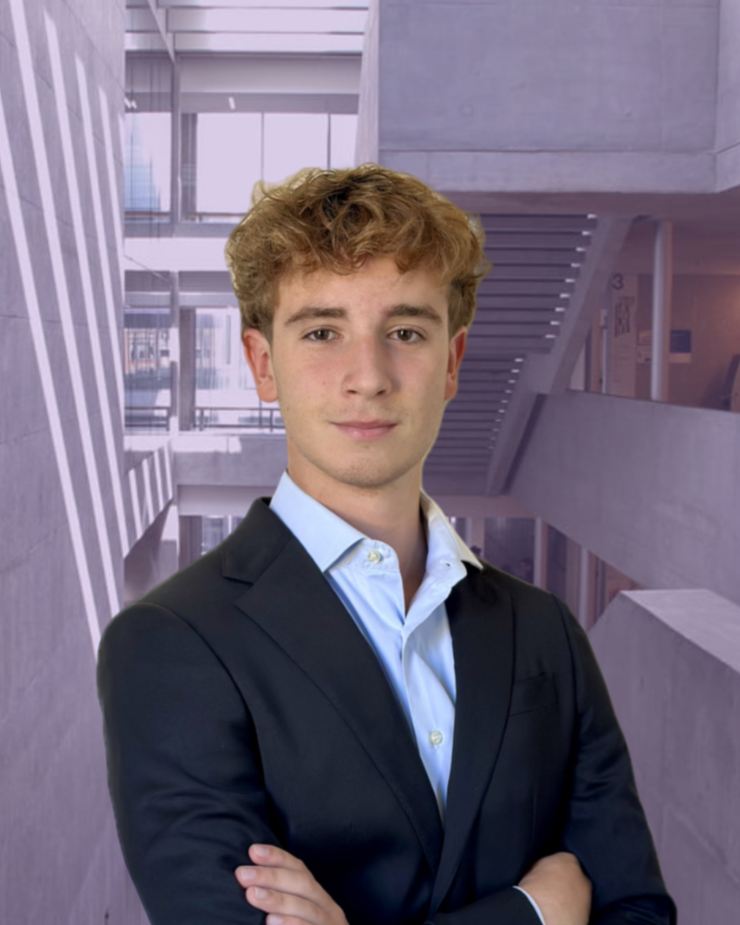 A person in a dark suit and light blue shirt standing outdoors in front of modern buildings.