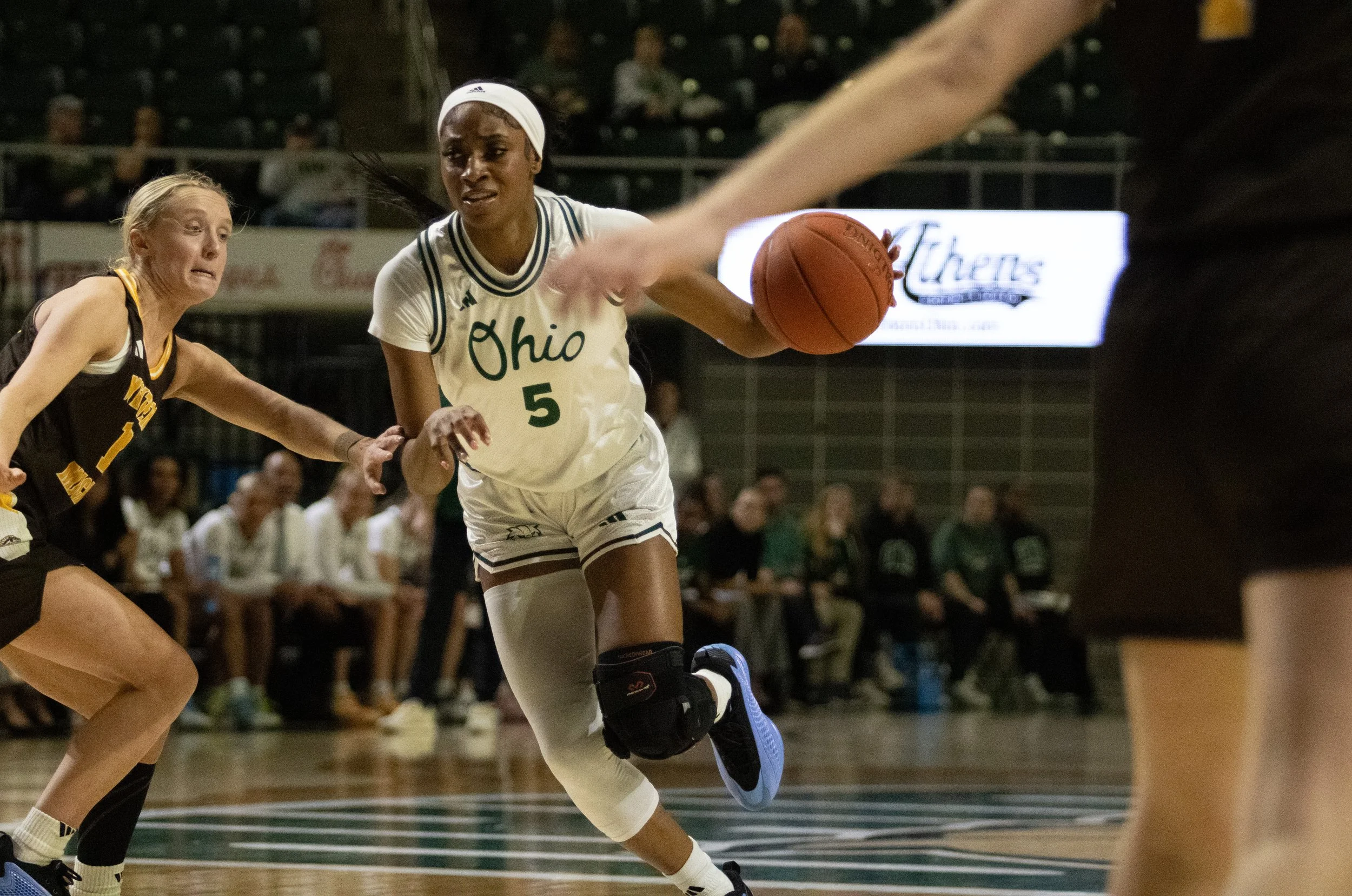 A female basketball player in a white Ohio uniform, number 5, dribbling the ball as she runs down the court during a game, with opponents and audience in the background.