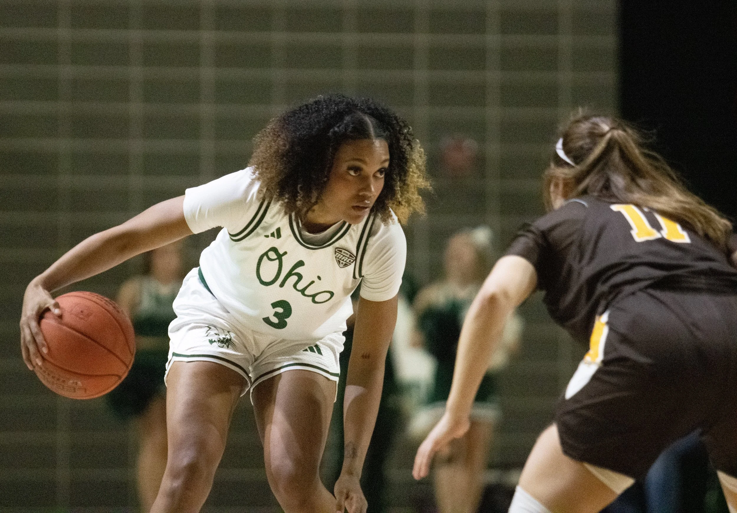 A female basketball player from Ohio wearing a white uniform with number 3, dribbling the ball and guarded by an opponent in a black uniform with the number 11, during a game.