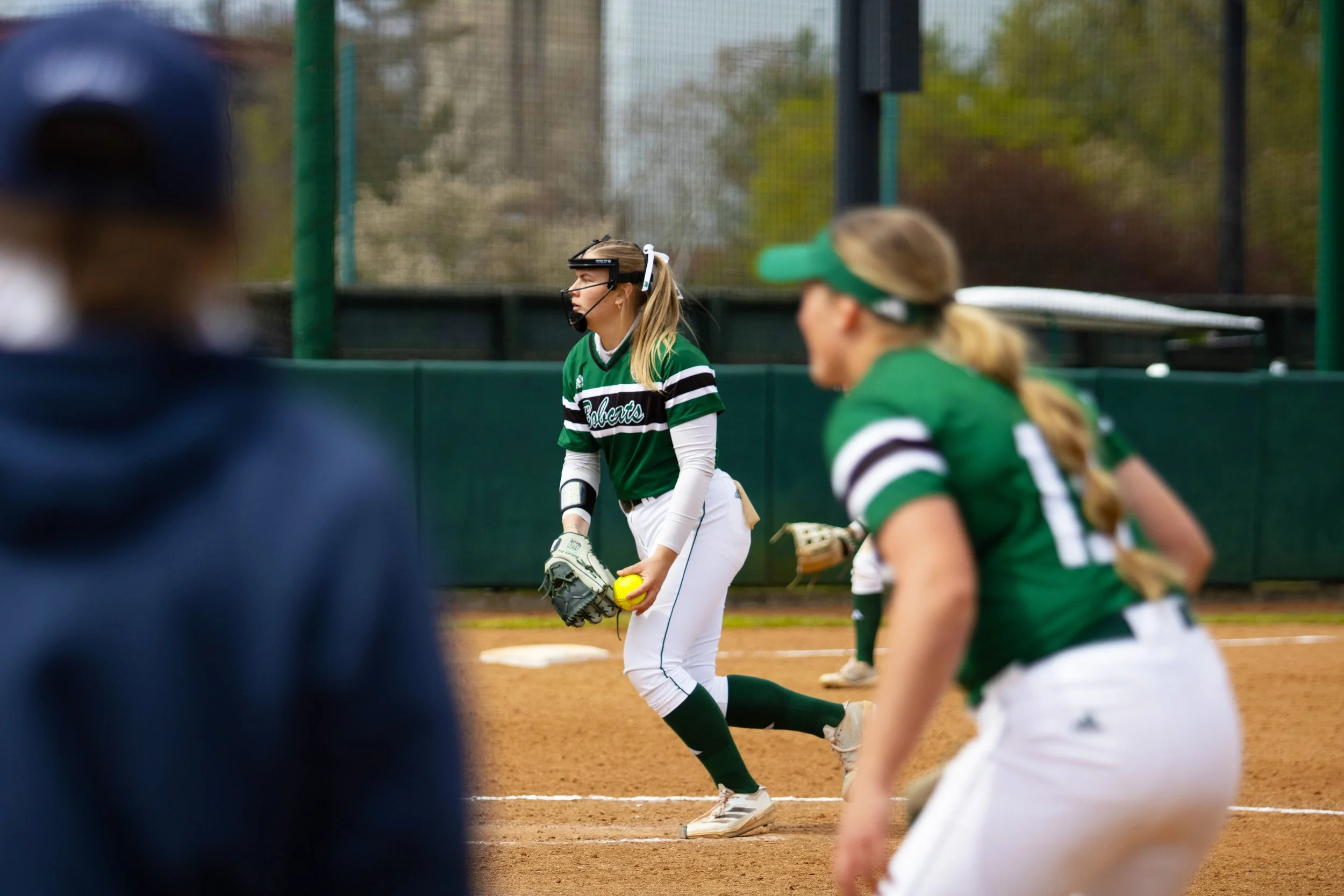 Softball players in green uniforms on a softball field, with one player preparing to catch a ball.