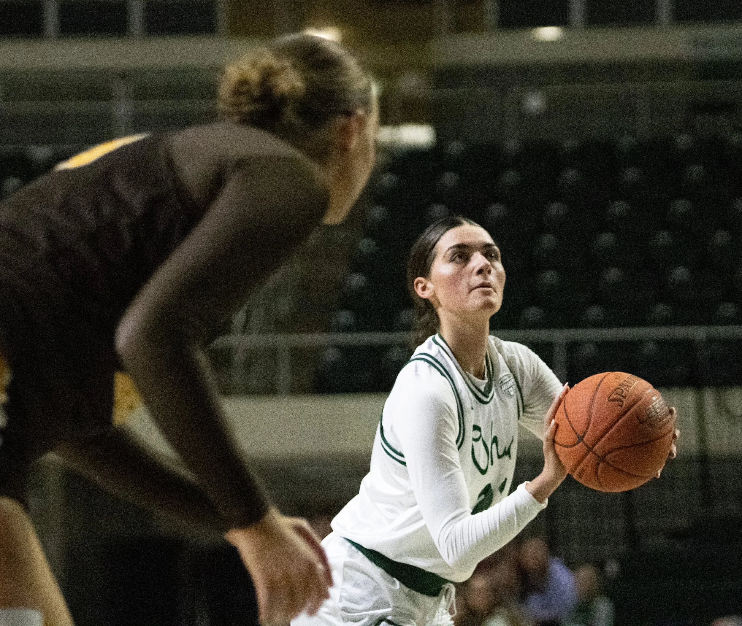 A female basketball player in a white uniform holding a basketball, looking up, while a woman in dark athletic attire crouches nearby during a game inside a gymnasium.