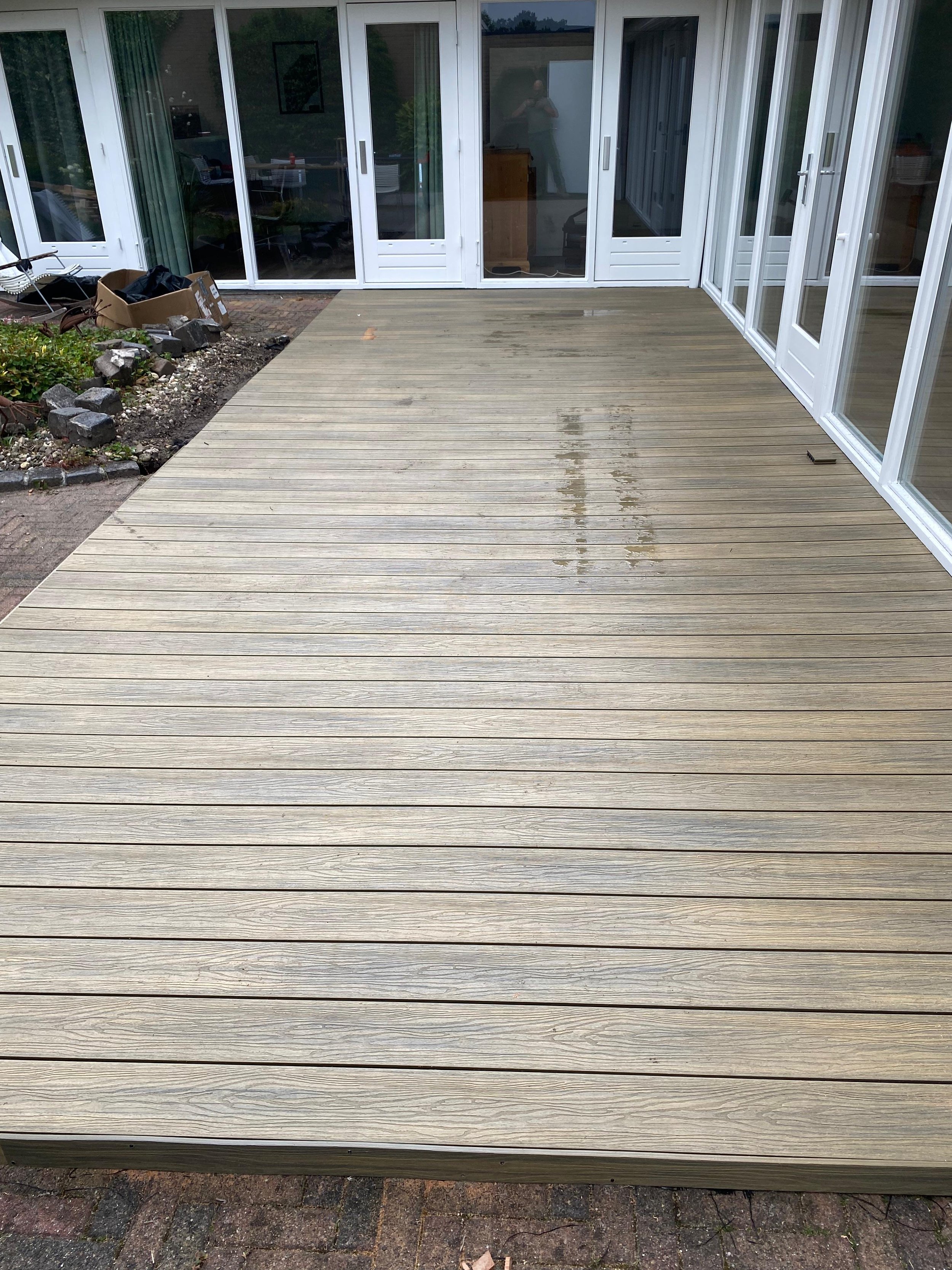 Gloednieuwe houten veranda 's achtertuin, waarvan het oppervlak nog nat is, naast een woning met grote glazen schuifdeuren.