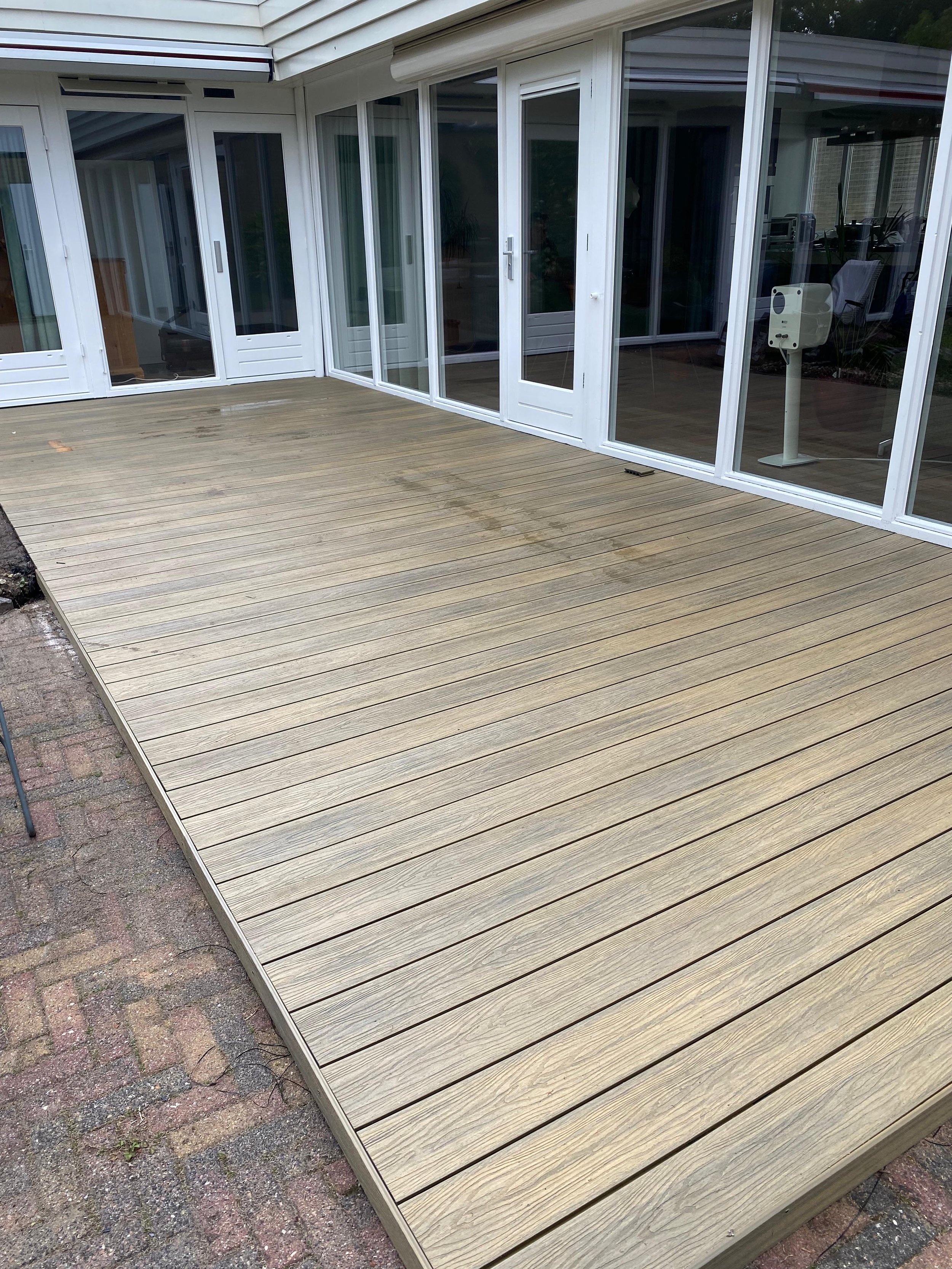 Buitenterras met houten vloer en grote glazen schuifdeuren van wit frame, kijkt uit op het interieur van een huis.