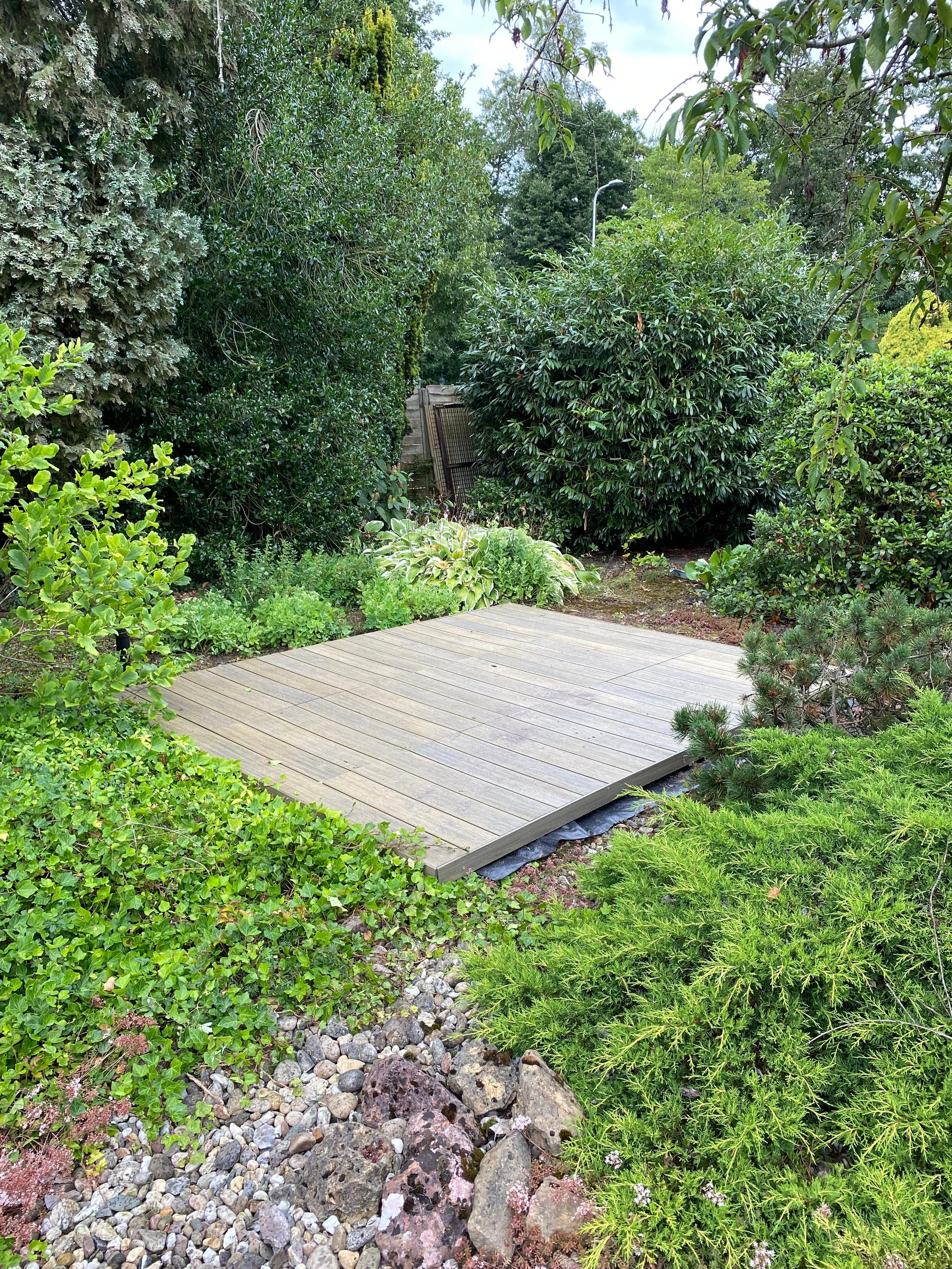 Tuin met houten terras omringd door groene struiken en bomen.