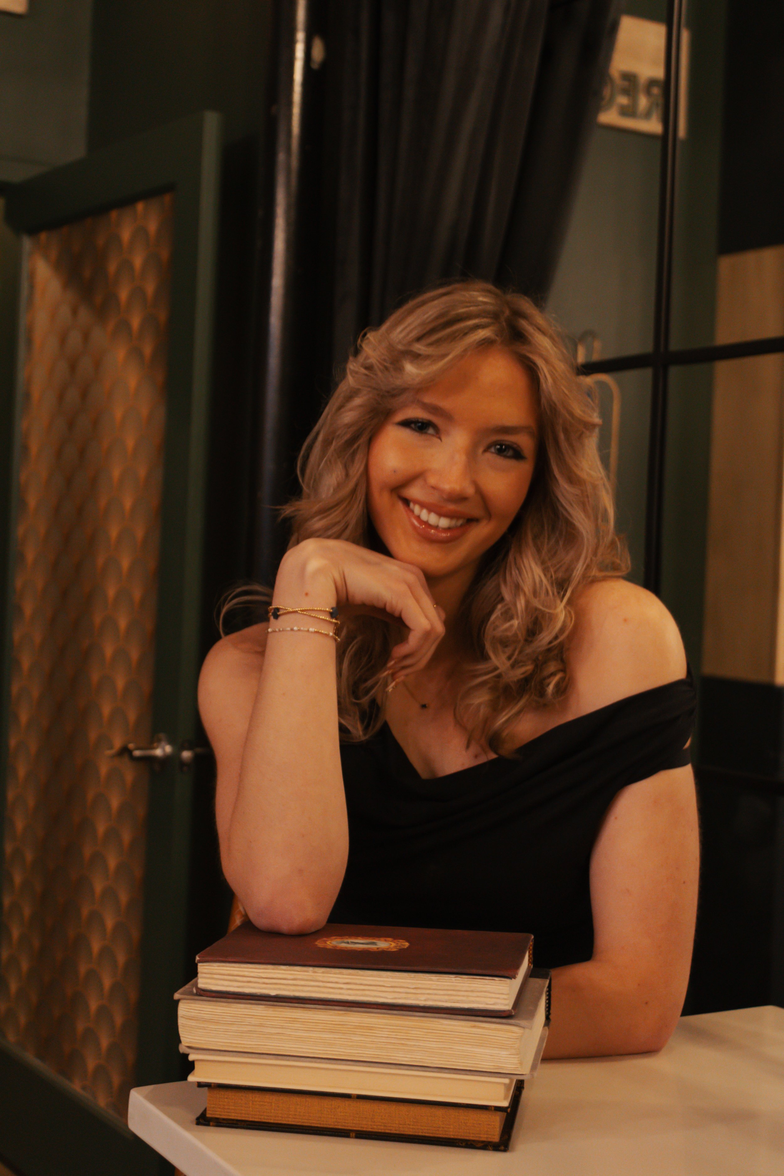 A woman with wavy blonde hair smiling, resting her chin on her hand, sitting at a table with a stack of books.