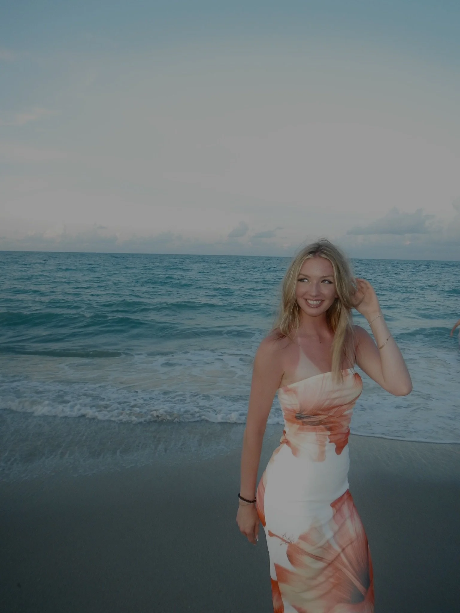 A young woman with blonde hair smiling at the beach with the ocean and sky in the background, wearing a strapless dress with orange and white patterns.