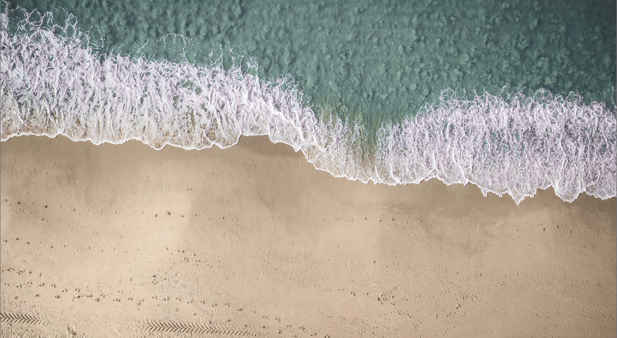 Aerial view of a sandy beach with gentle ocean waves washing onto the shore, footprints and tire tracks visible in the sand.