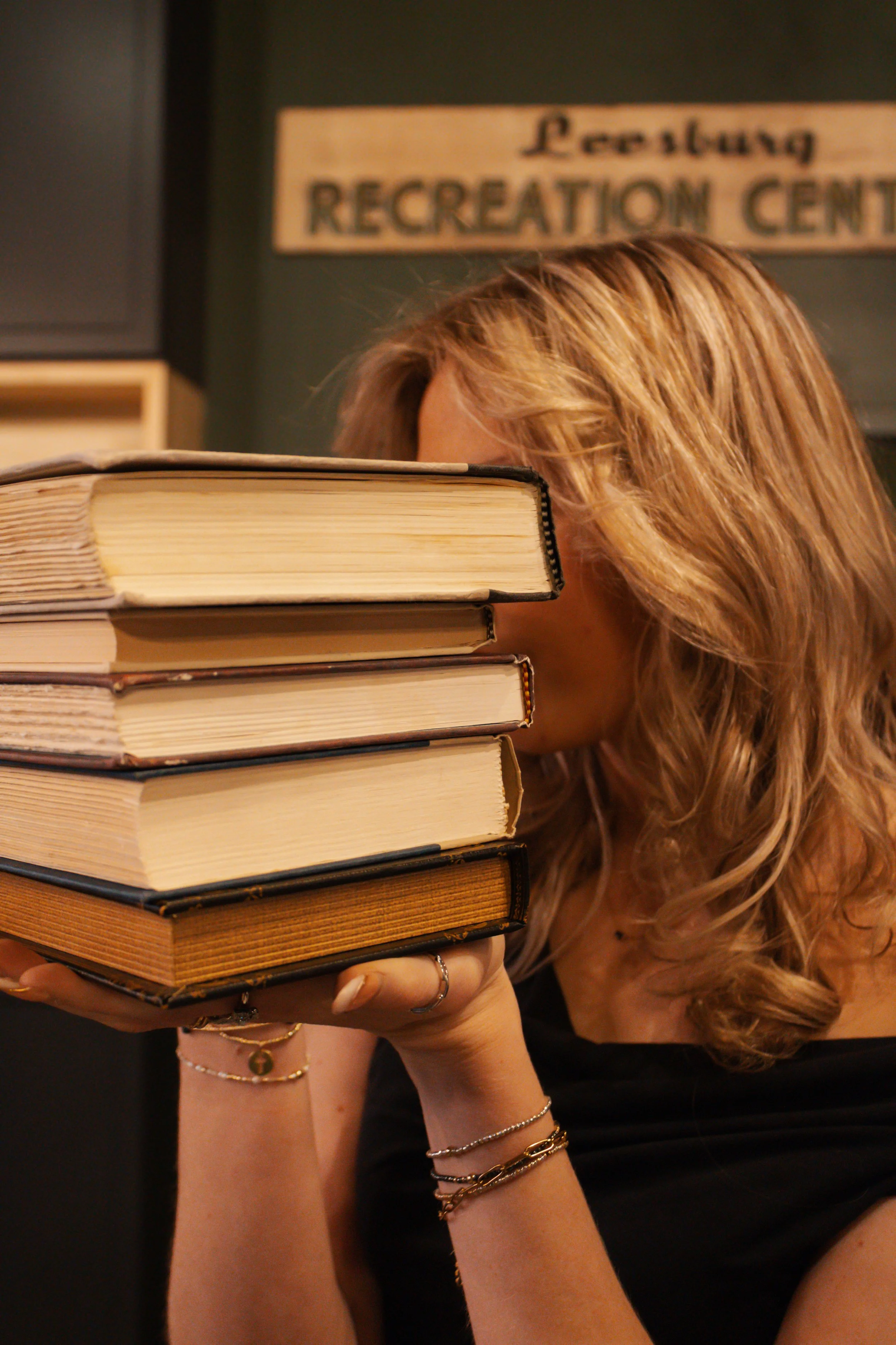 A woman with wavy hair covering part of her face, holding a stack of books, in front of a sign that reads 'Recreation Center'.