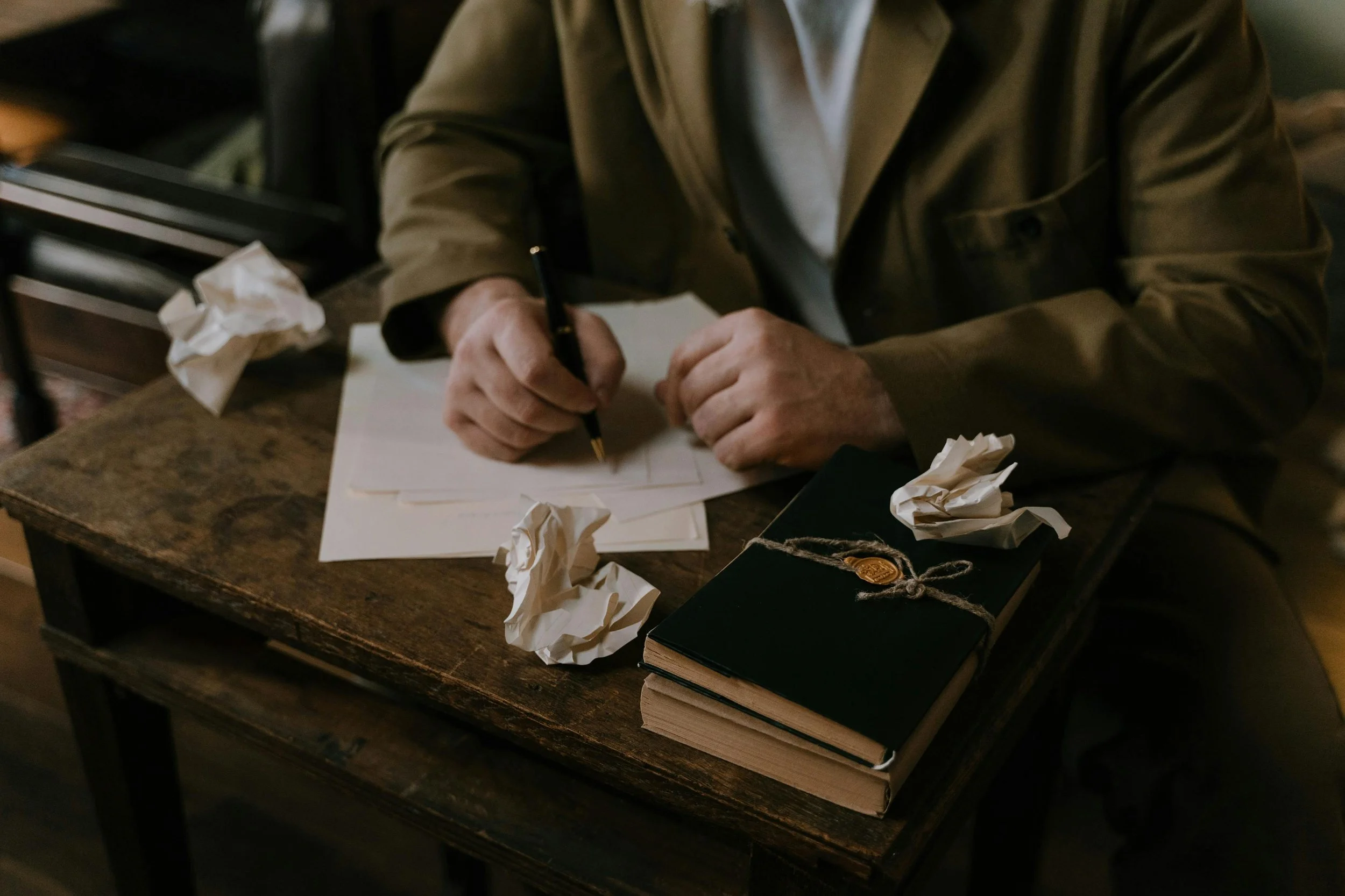 A person in a brown jacket sitting at a wooden desk, writing on a piece of paper with crumpled papers scattered around, and a stack of black books secured with twine and a wax seal.