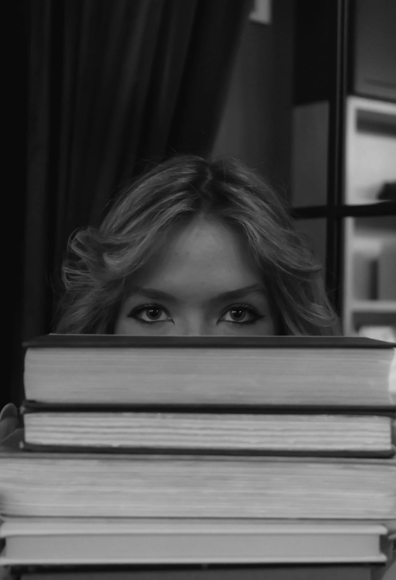 Person with styled hair peeking over a stack of books, eyes visible above the books.